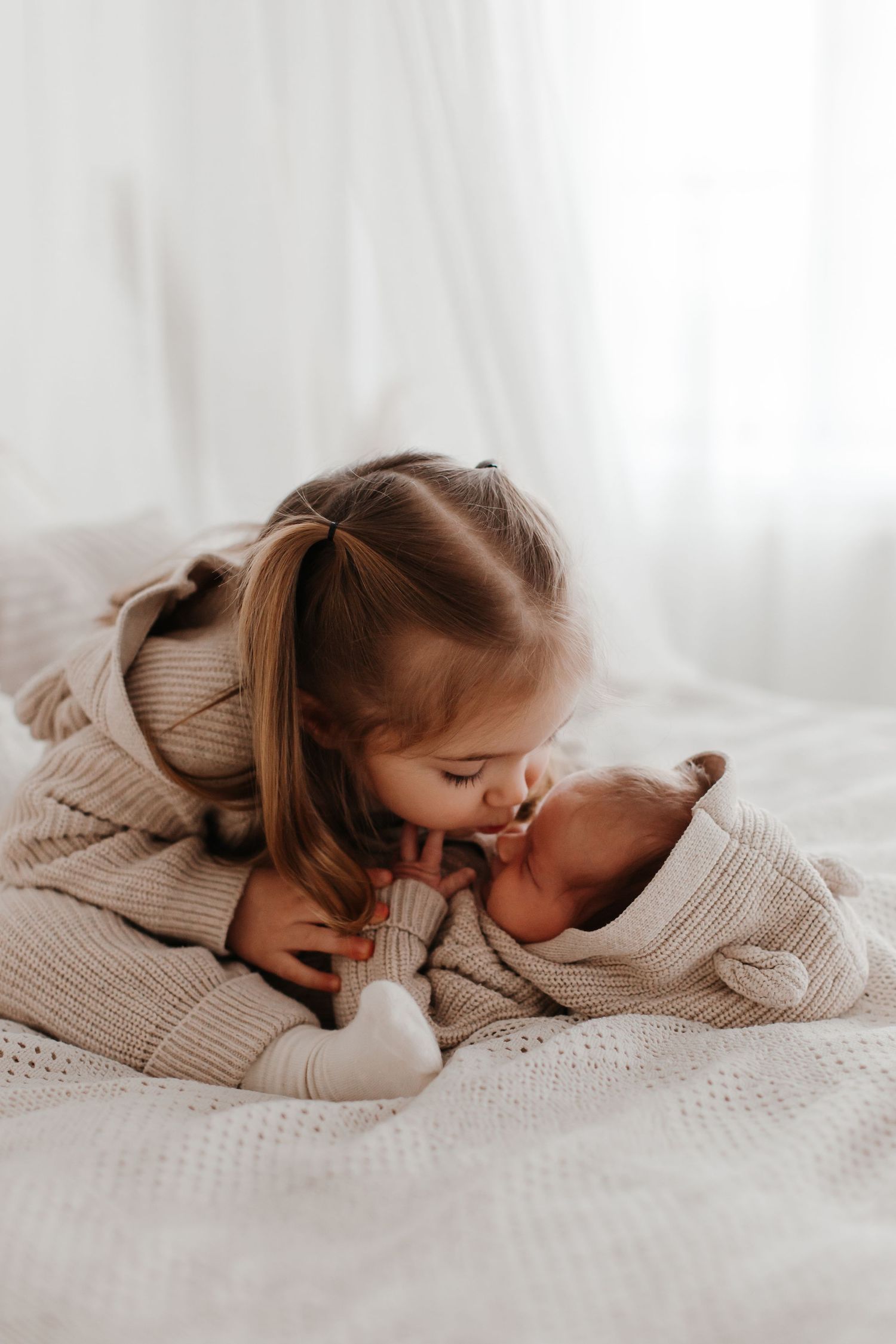 Young child in sweater gently kisses newborn sibling on white bedding.