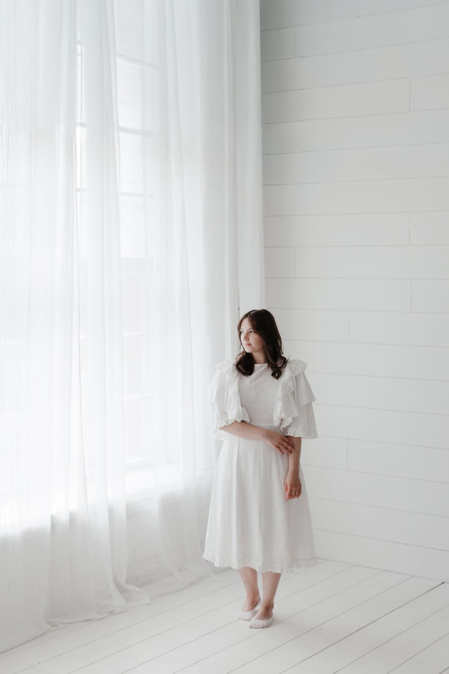 People in white dresses stand against white wall in bright minimalist setting.
