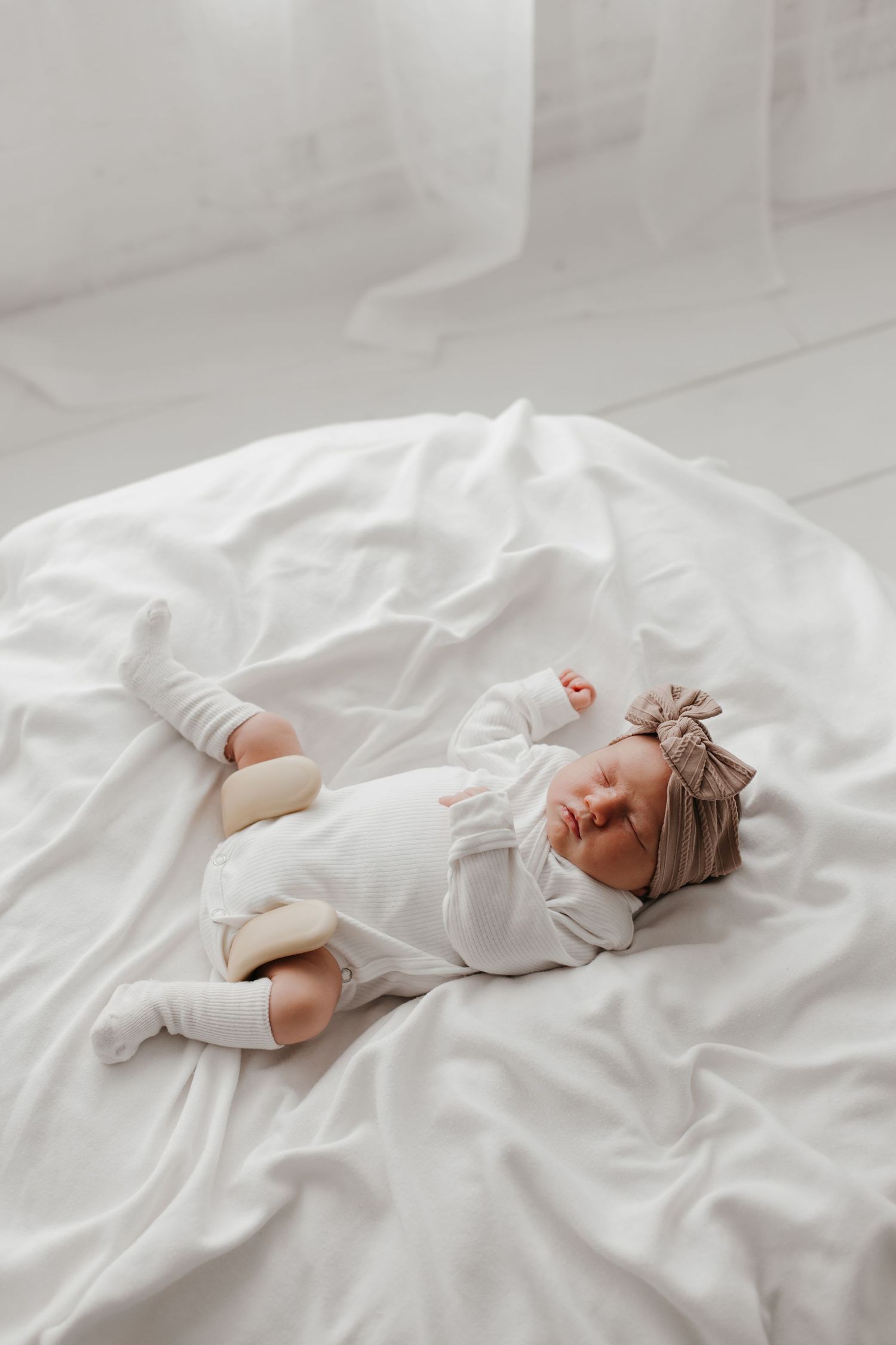 Sleeping newborn in white onesie lies peacefully on white bedding.