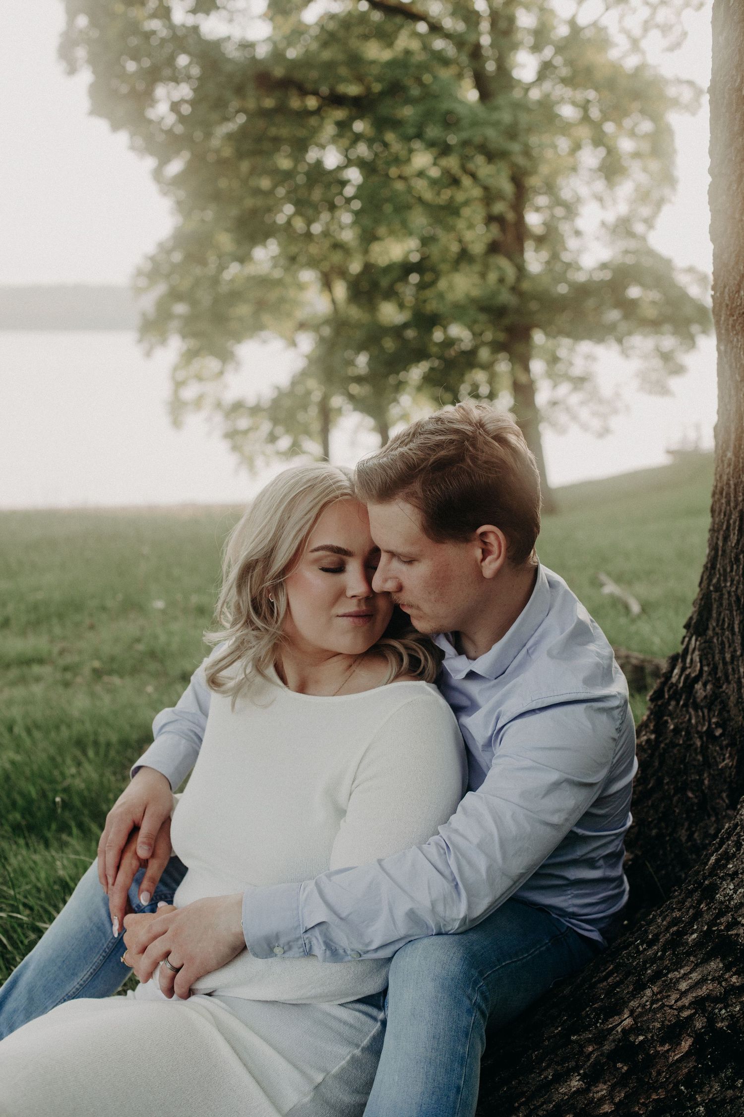 Couple sitting against a tree by a lake, sharing a tender moment at sunset.