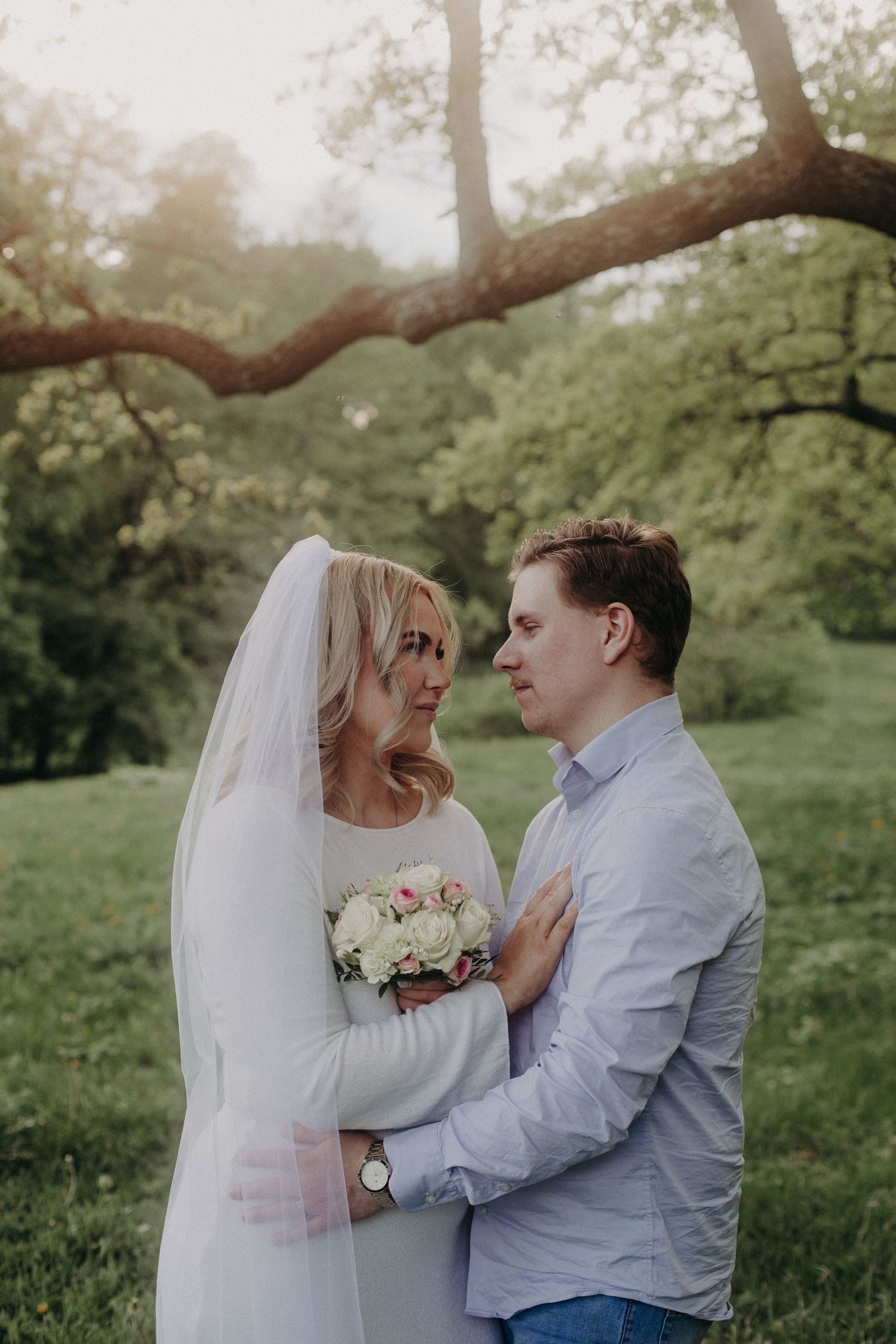 Bride in white dress and veil holds bouquet while gazing at groom in blue shirt outdoors.