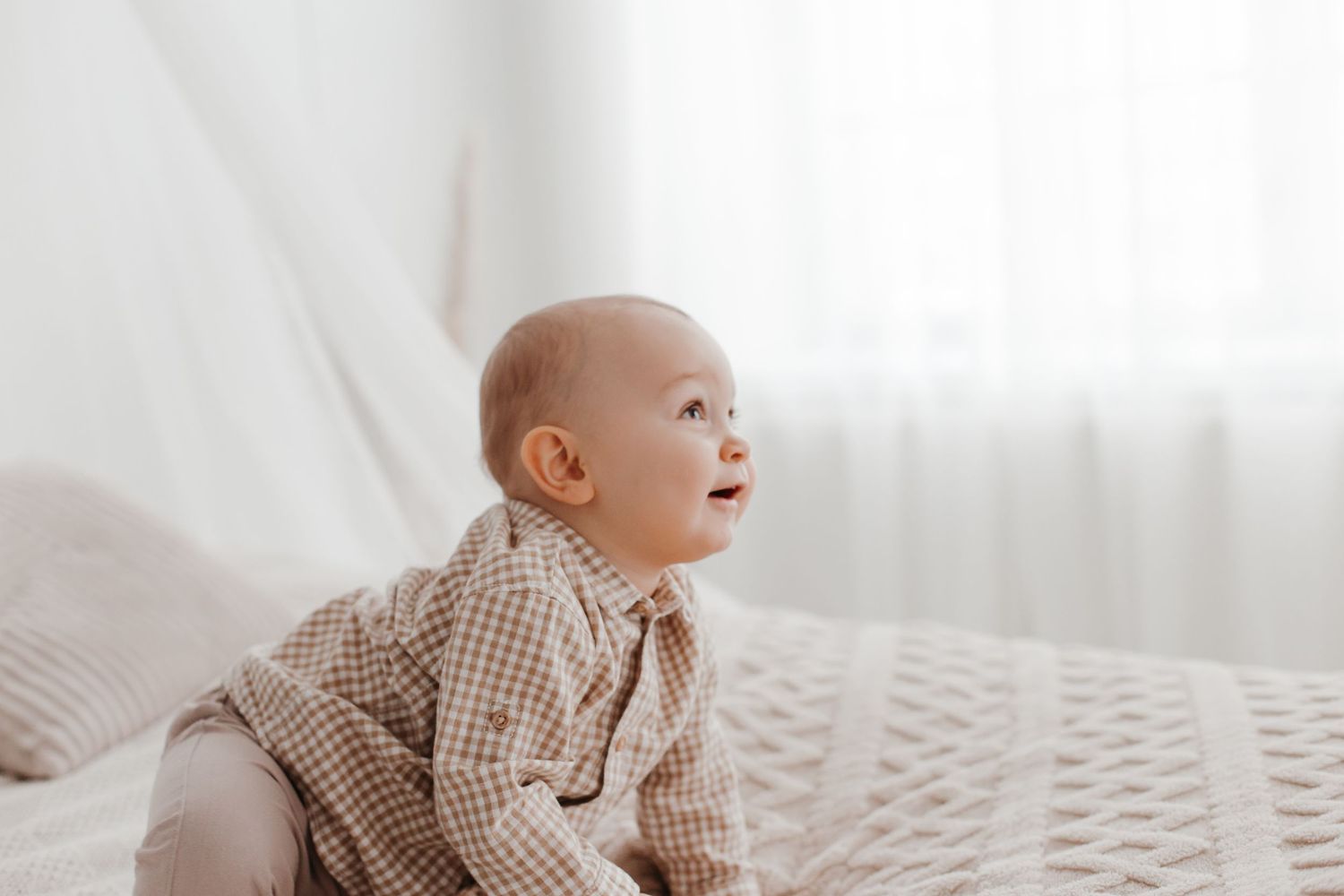 Baby in checkered shirt looking up while sitting on textured white bedding.
