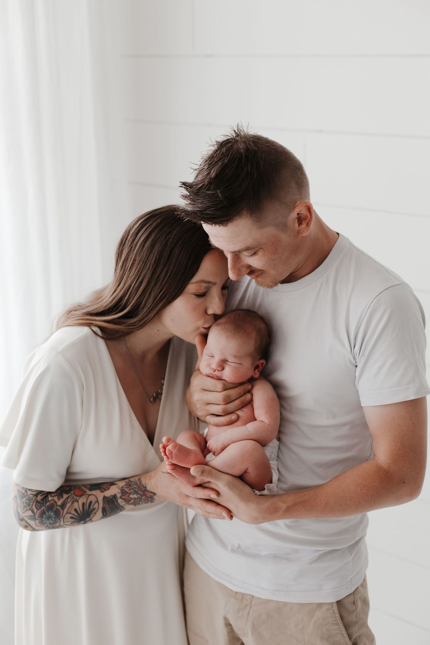Parents sharing tender moment with newborn baby against white wall.