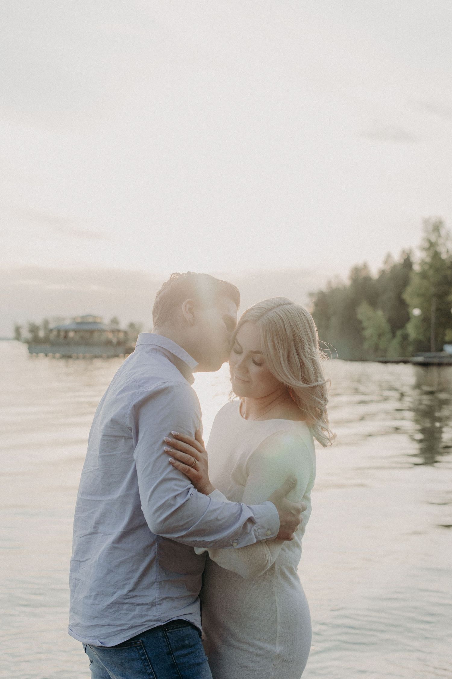 A couple shares a tender kiss by a sunlit lakeside, the woman in a white dress embraced by a man in a light blue shirt.