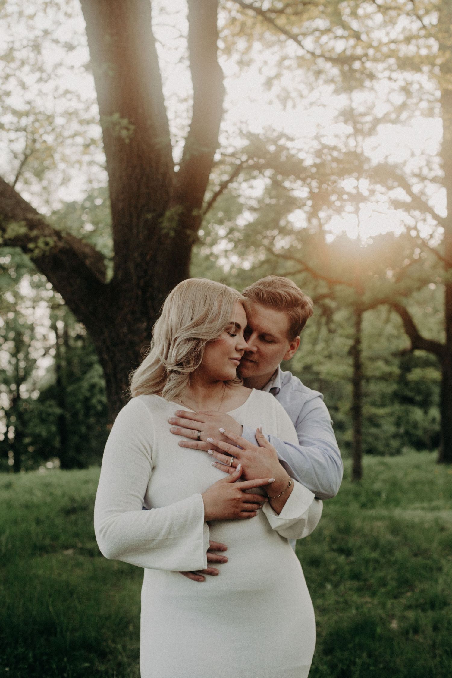 Groom tenderly embraces bride in white dress from behind near sunlit trees.
