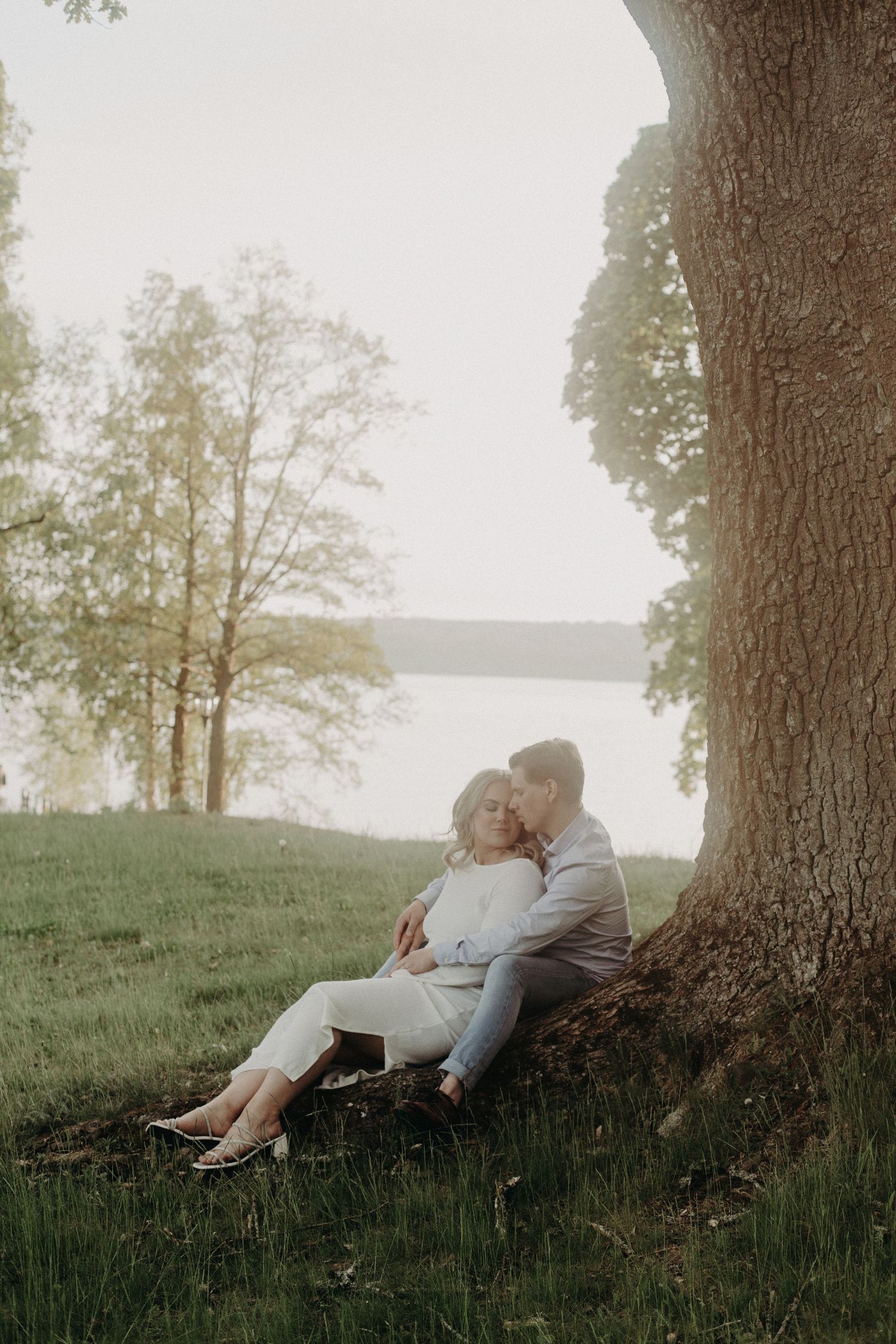 Couple sitting together against a large tree by a lake in a lush green park during golden hour.