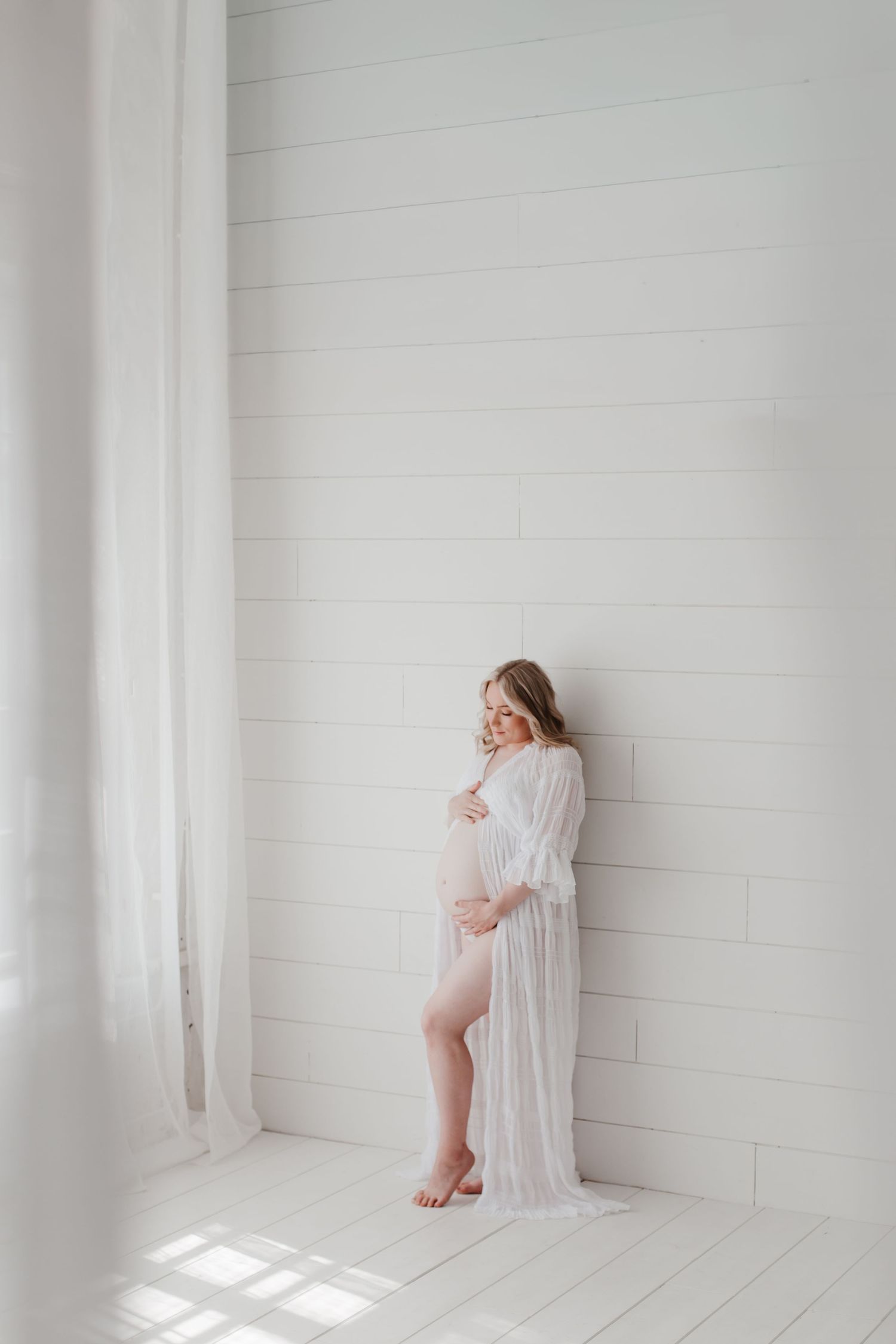 Artistic maternity photograph with soft window light streaming across white room.