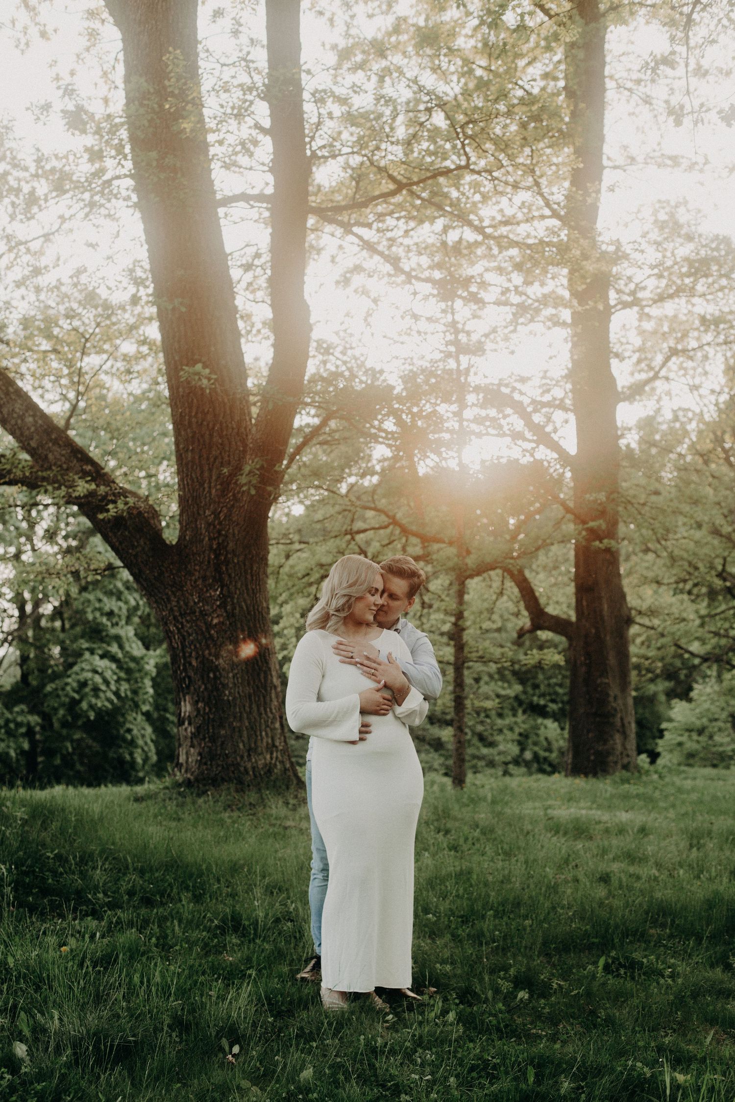 Couple embracing in sunlit forest, pregnant woman in white dress, golden hour light filtering through trees.