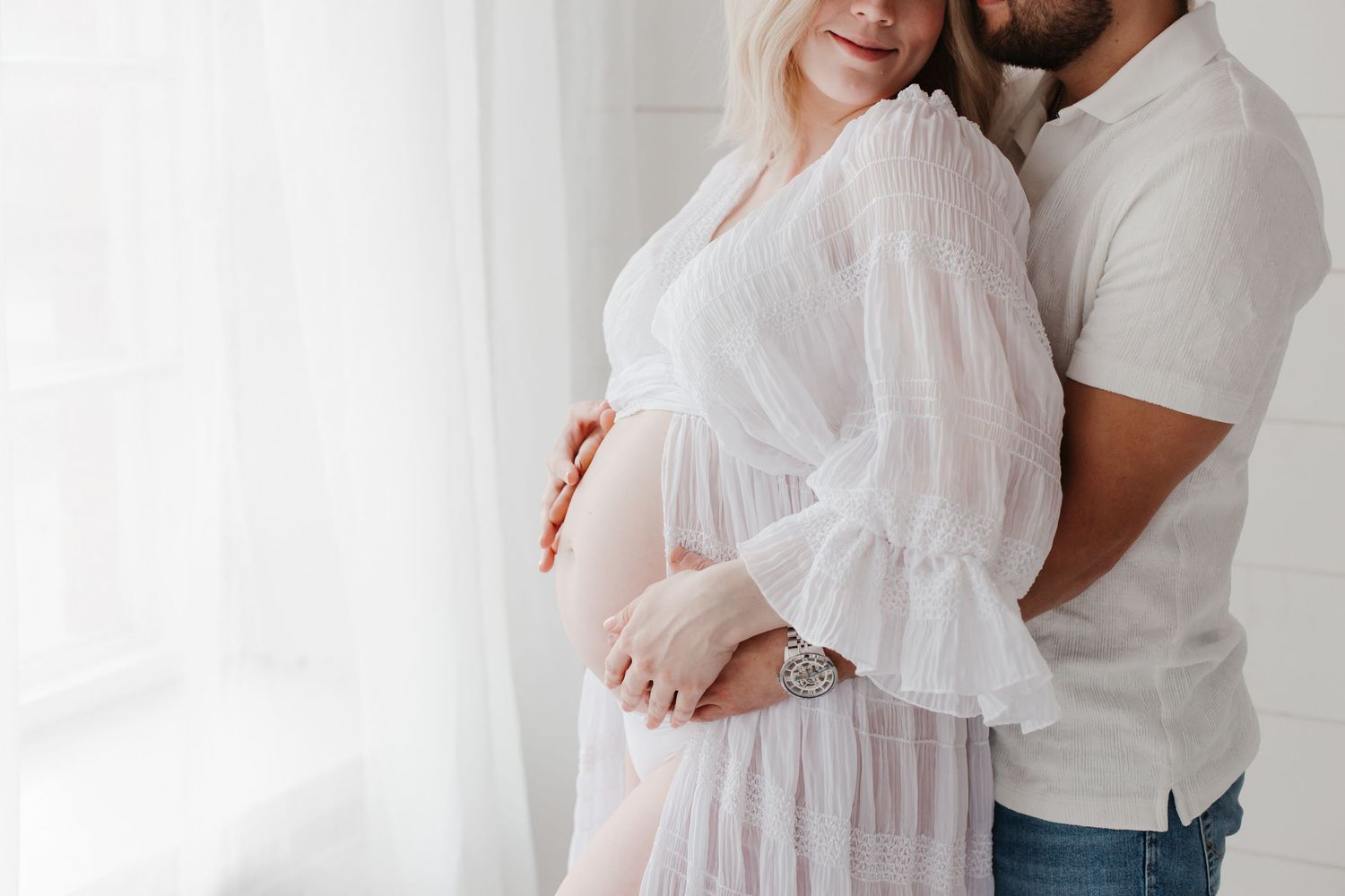 Couple shares tender moment during maternity photoshoot in bright white studio space.