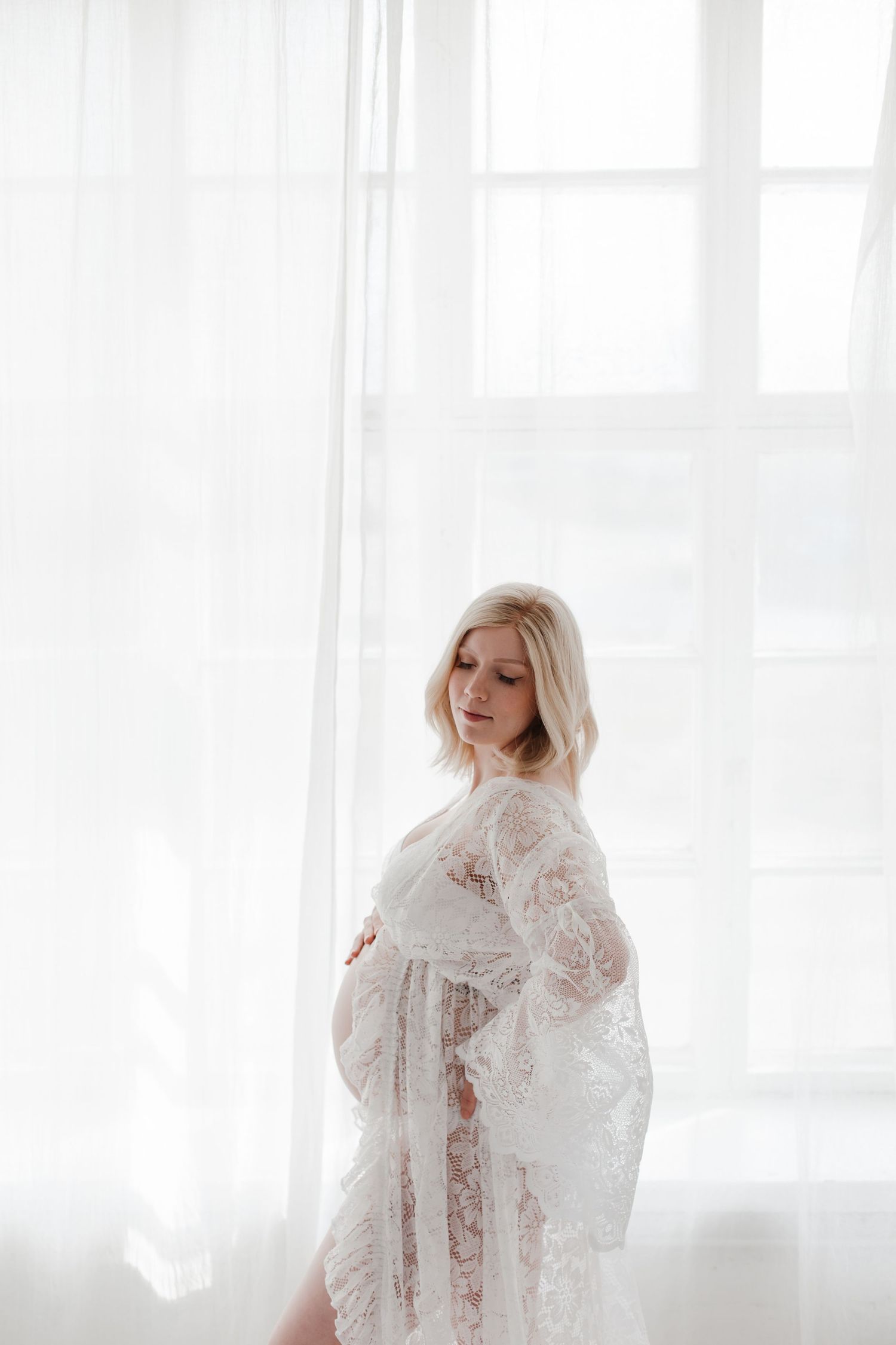 Dreamy maternity portrait with white lace dress against bright window light.