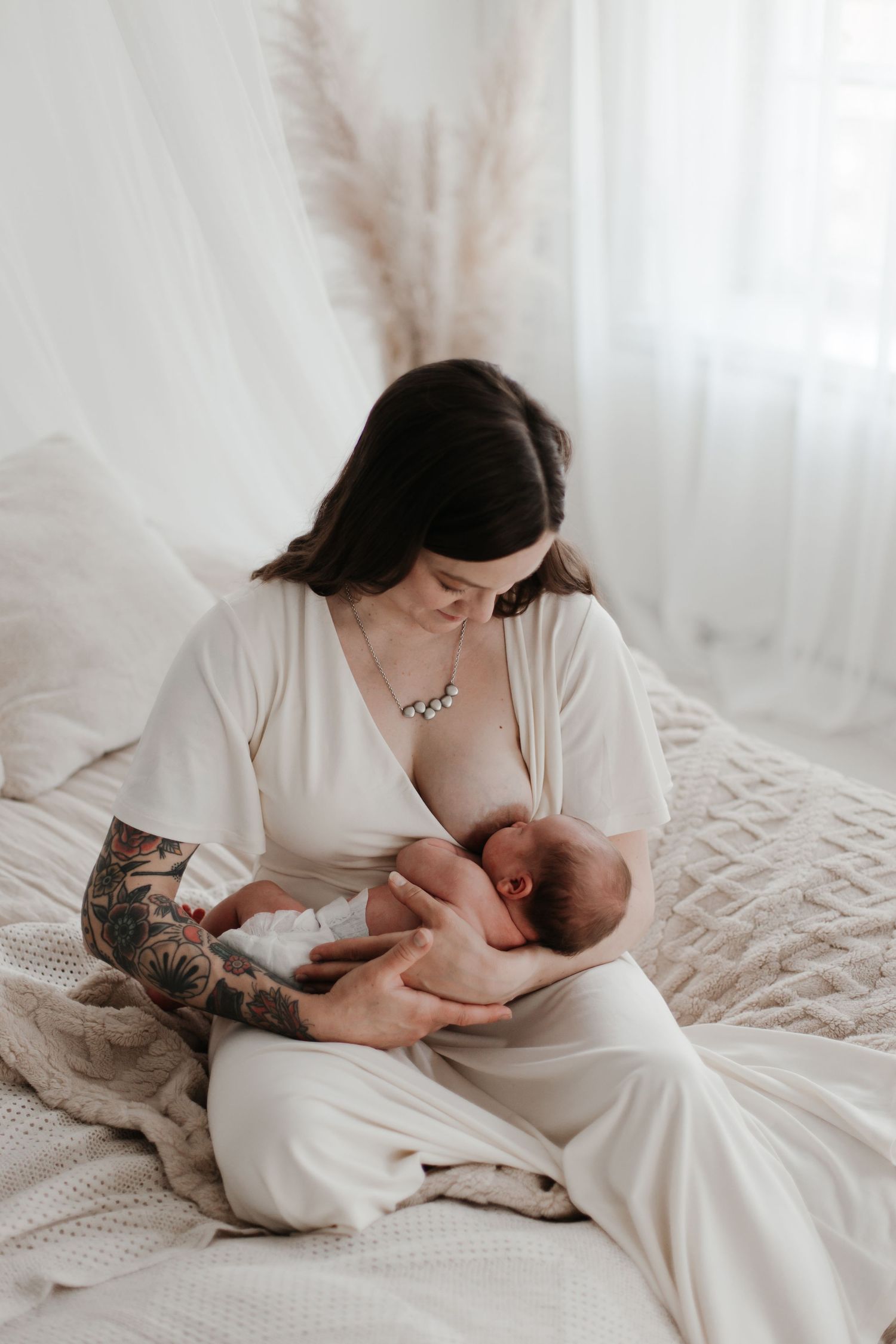 Mother in white dress nursing newborn baby while resting on white bedding.