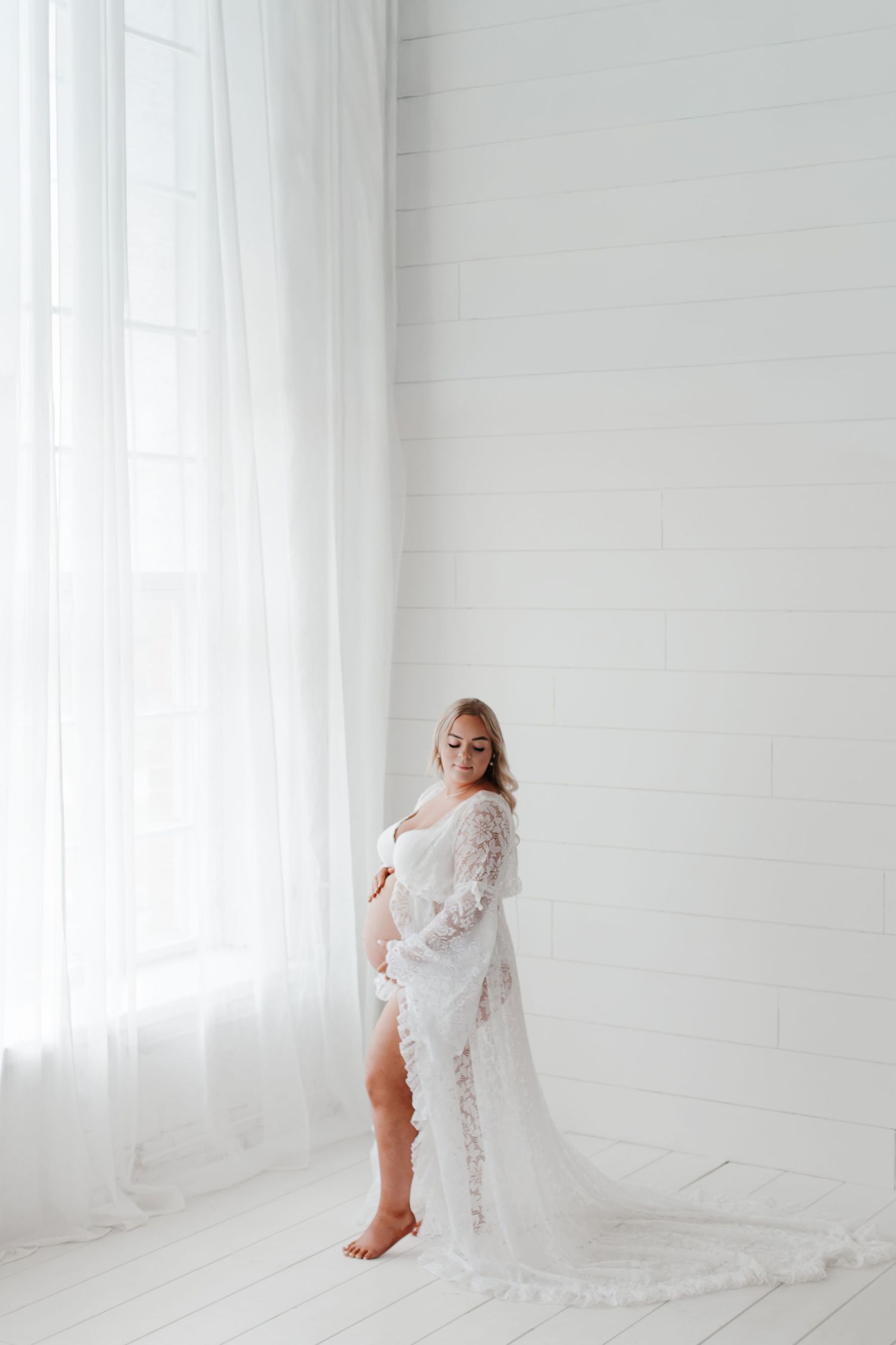 Maternity photo featuring white lace gown in bright minimalist setting.