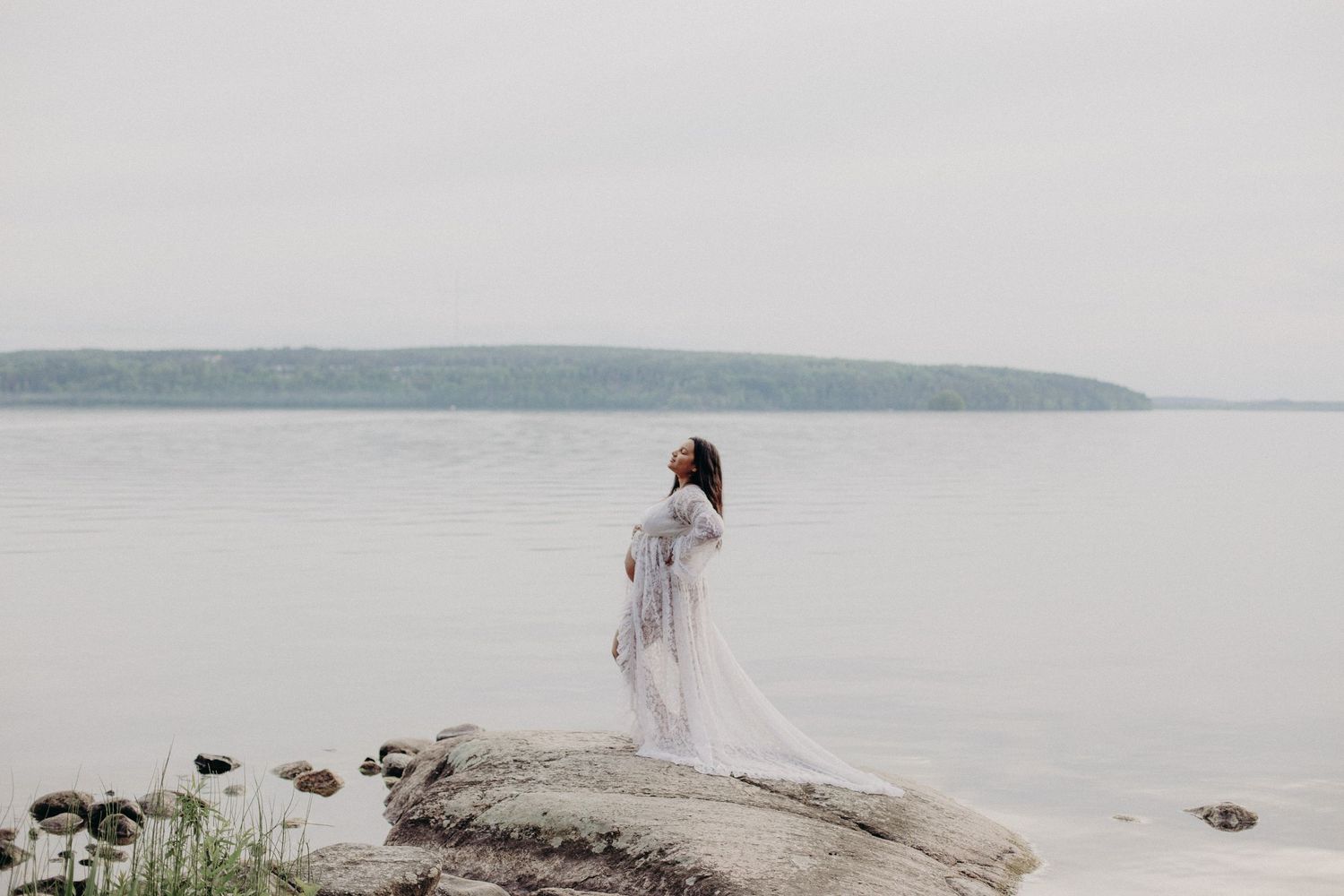 A pregnant woman in a flowing white gown stands on rocky shoreline with calm water and misty hills in the background.
