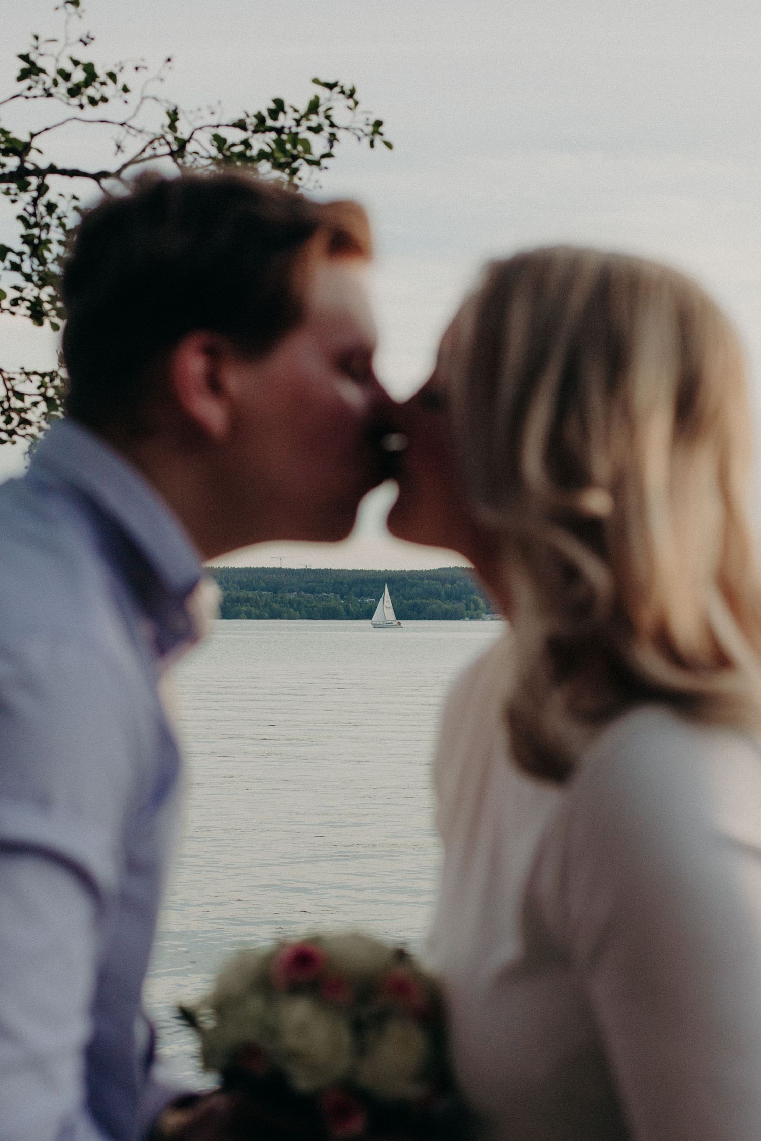 A couple shares a romantic kiss in front of a serene lake with a sailboat, the bride holding a bouquet of flowers.