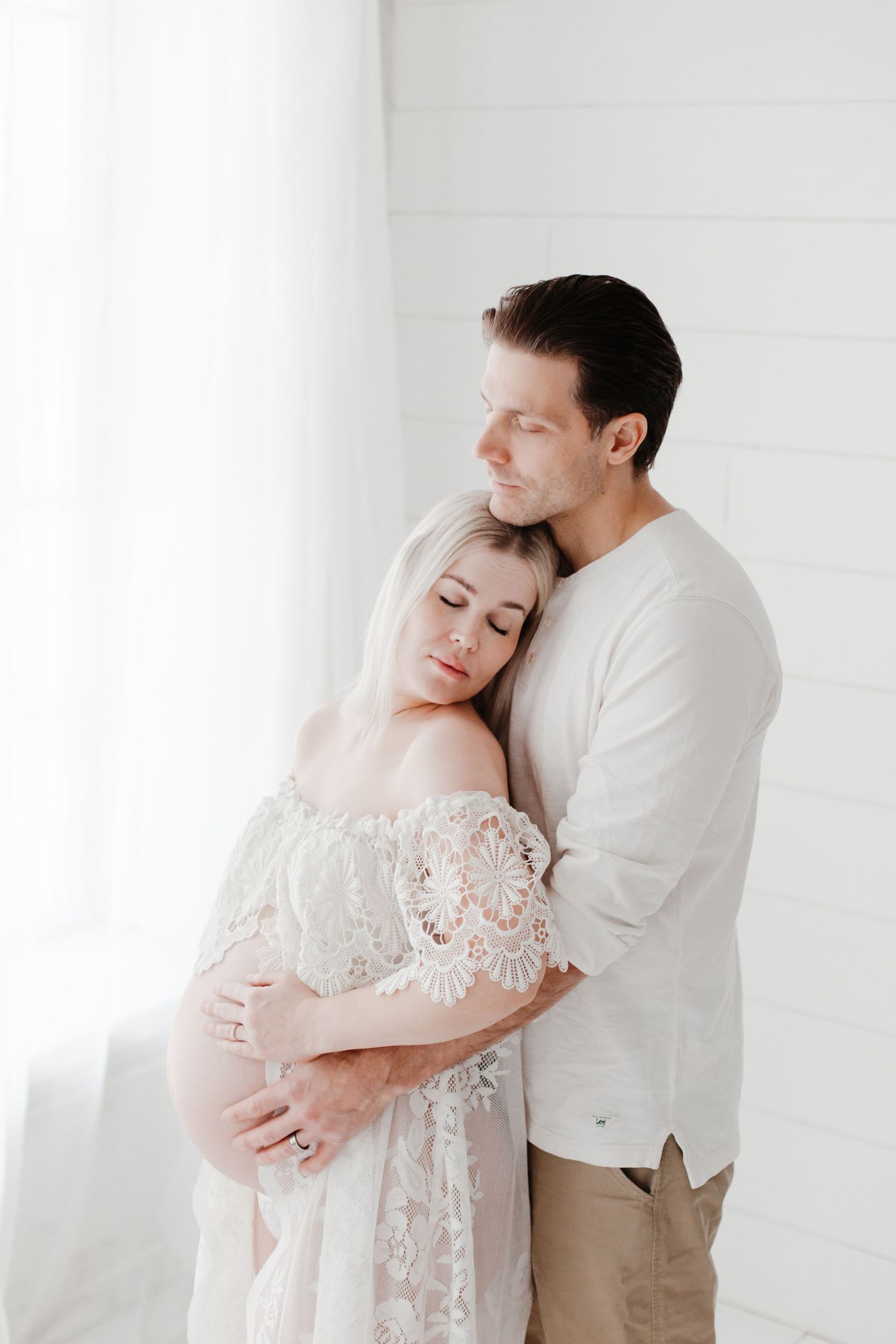 Couple in white embracing against white wall in intimate moment.