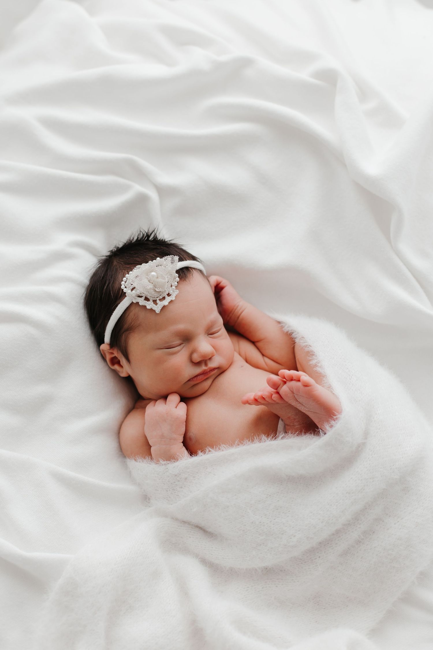 Sleeping newborn wearing delicate headband on white blanket.