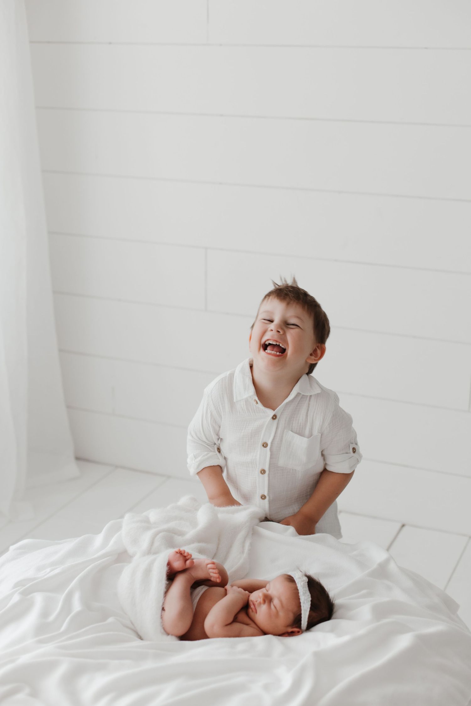 A baby in white clothing laughs joyfully while sitting on white bedding in a bright room.