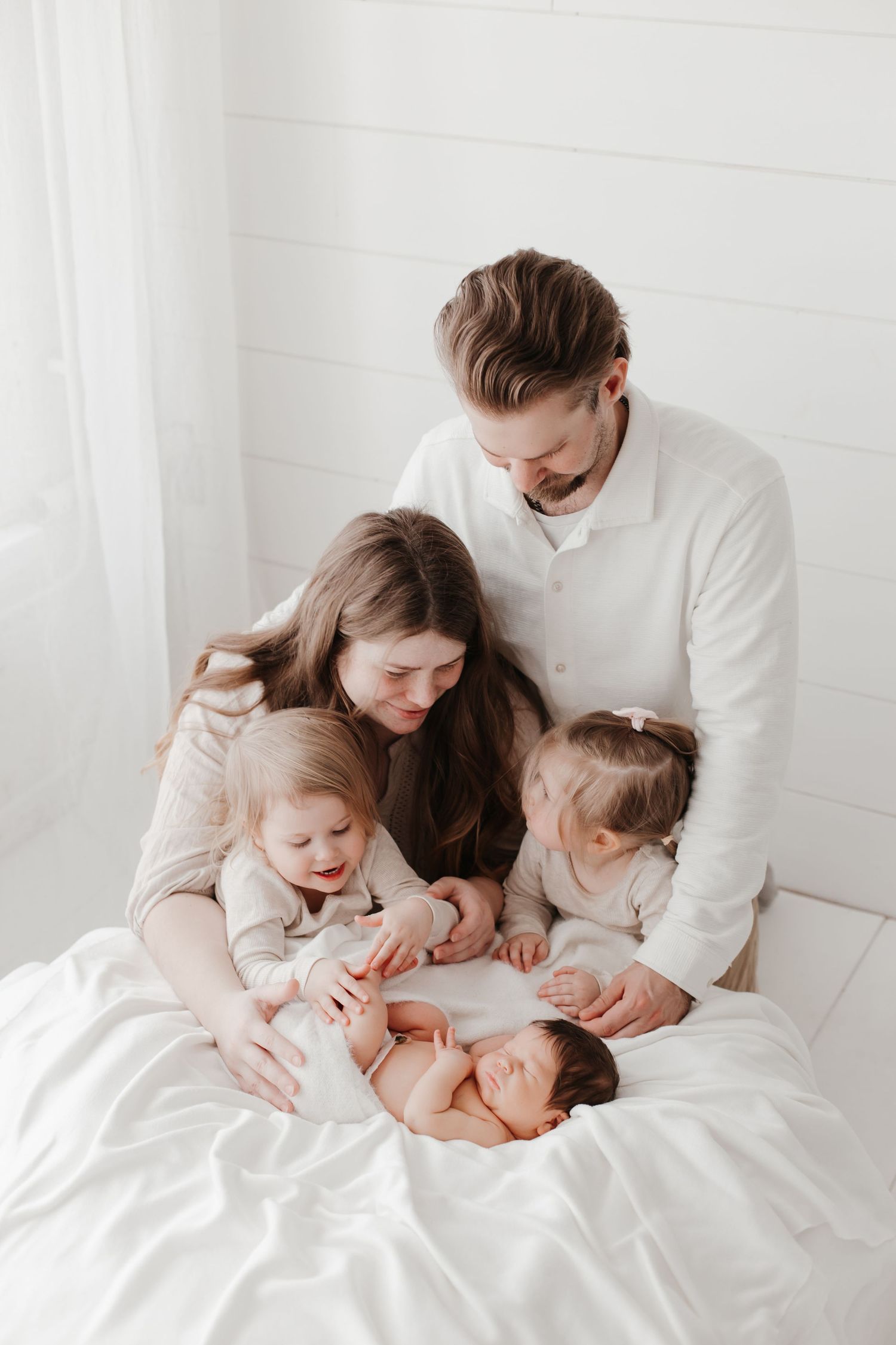 Family sharing intimate moment with newborn baby on white bedding in bright minimalist setting.