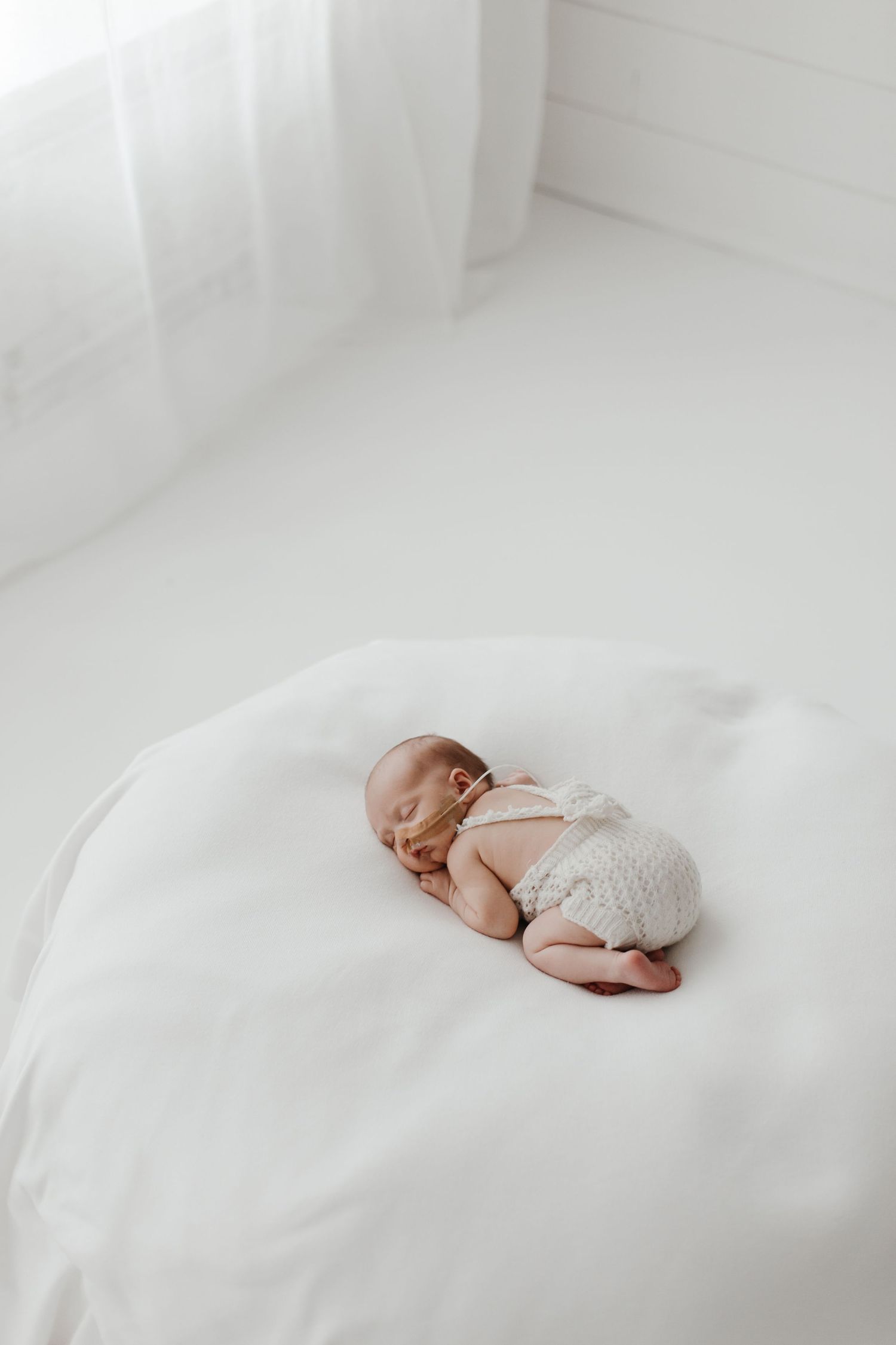Newborn in white onesie sleeping peacefully on white bedding.