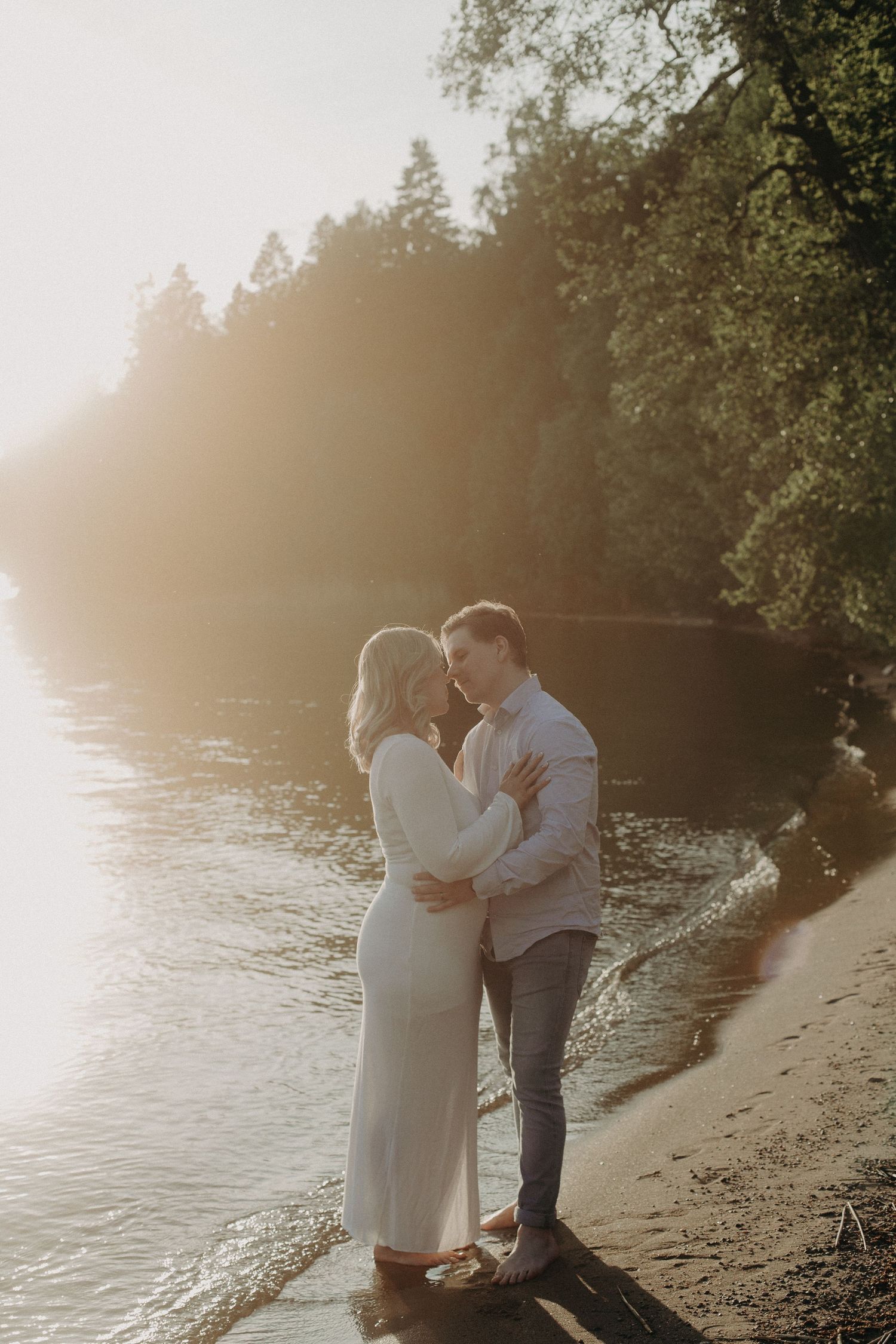 Couple embracing on a sandy riverbank at golden hour, surrounded by lush trees and soft glowing sunlight.