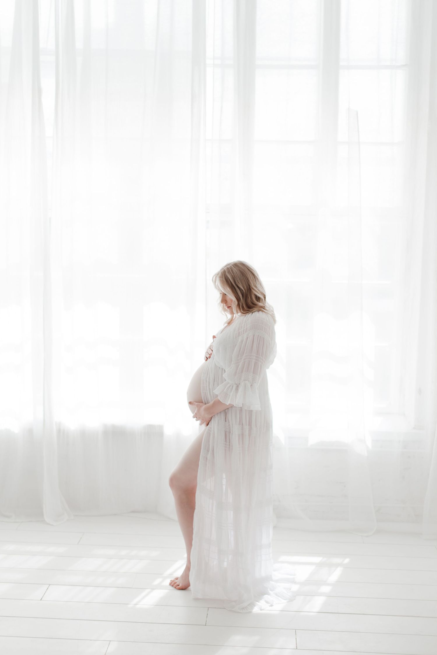 Dreamy maternity portrait in flowing white dress against bright minimalist backdrop.