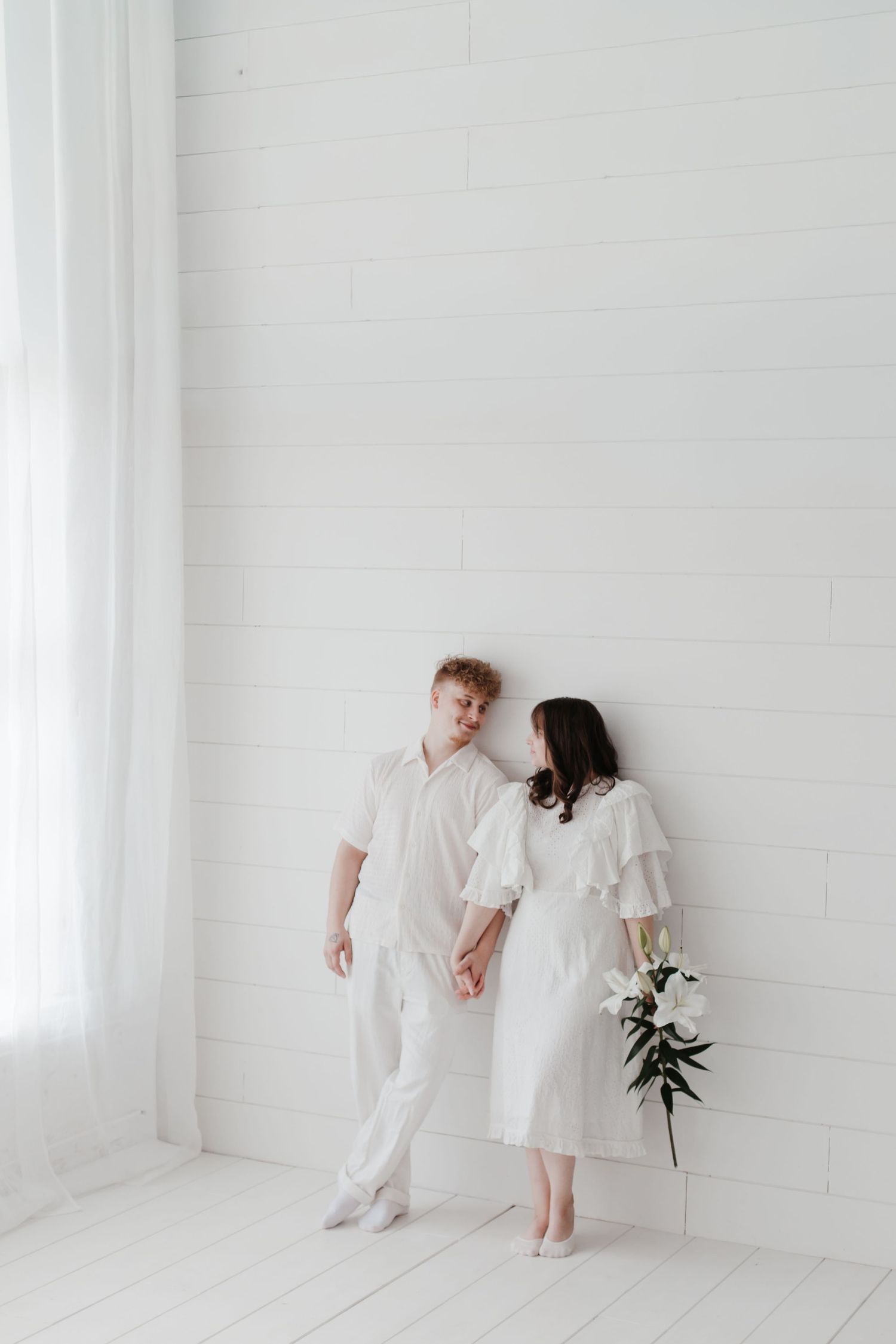 Family wearing white clothing poses together in a minimalist white room with natural lighting.