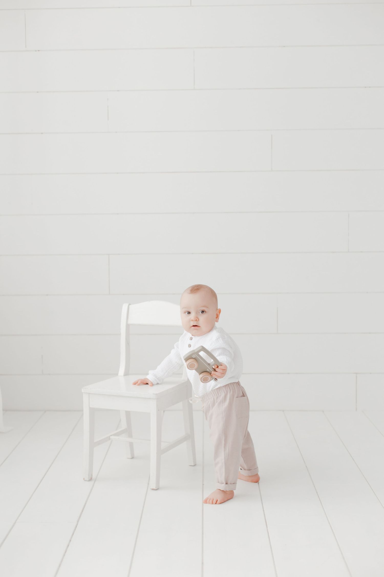 Small child in white shirt and beige pants standing beside white chair in minimalist setting.