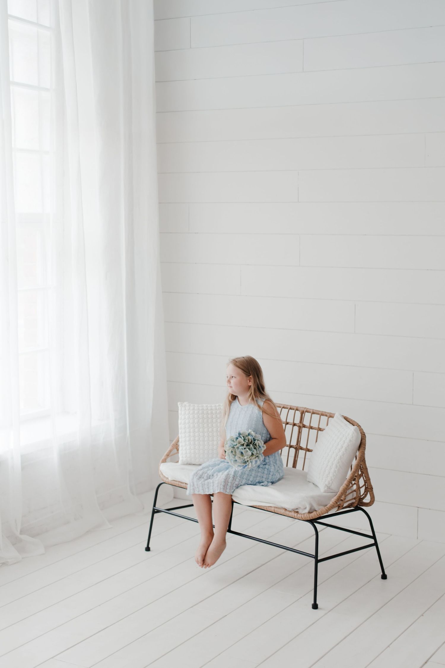 Child lounges on stylish rattan bench in light blue dress against white backdrop.