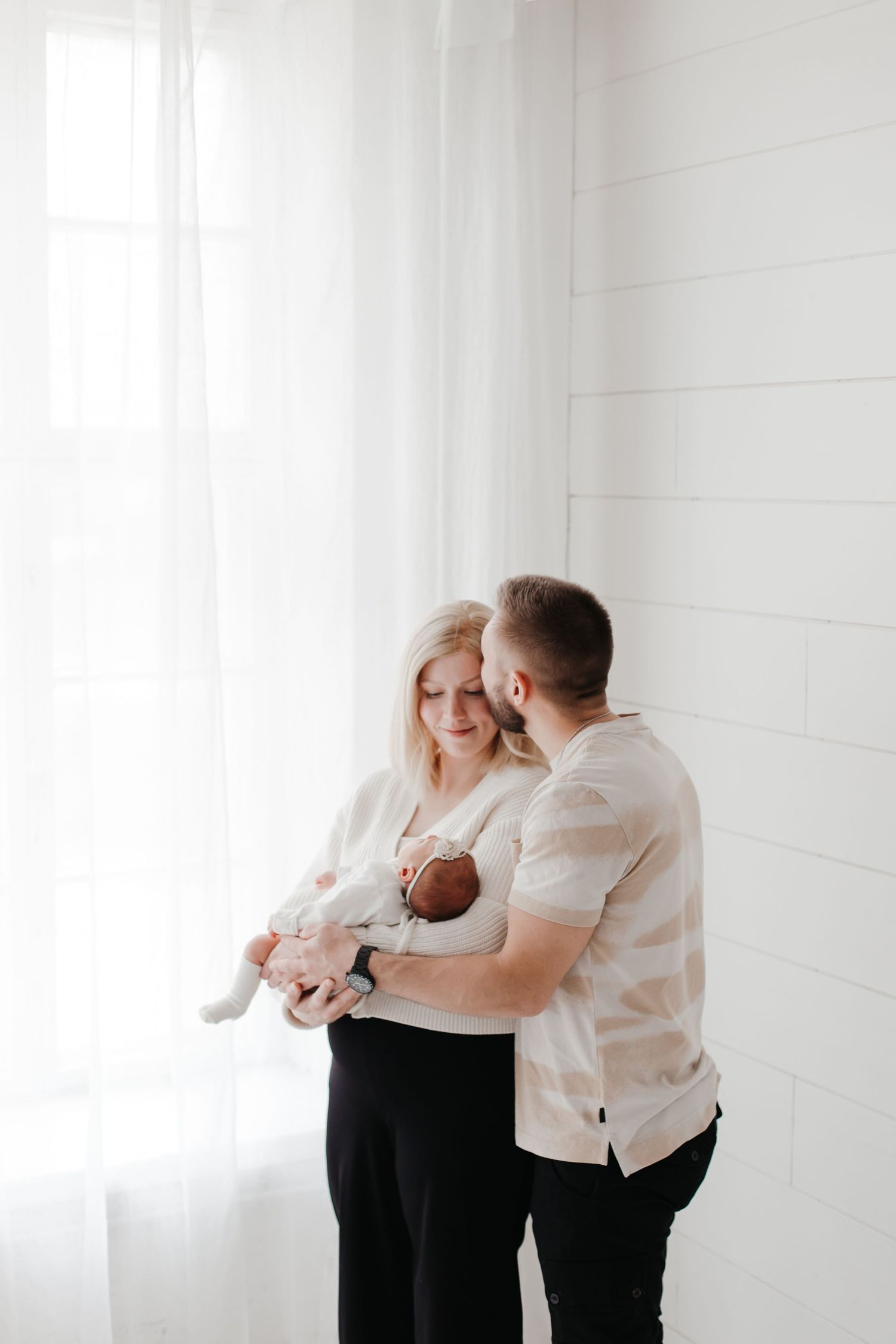 Parents sharing intimate moment with newborn baby by bright window.