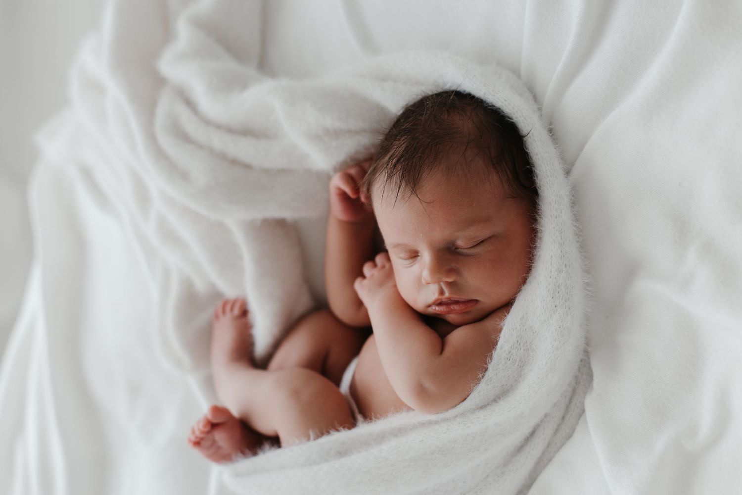 Sleeping newborn baby wrapped in white blankets in soft natural light.