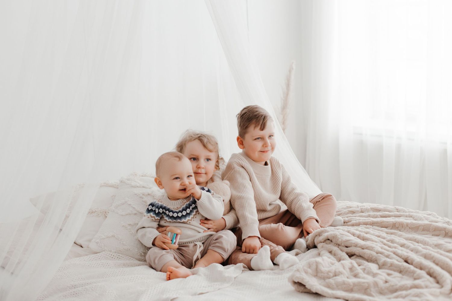 Three small children sitting together on white bed wearing neutral colored outfits.