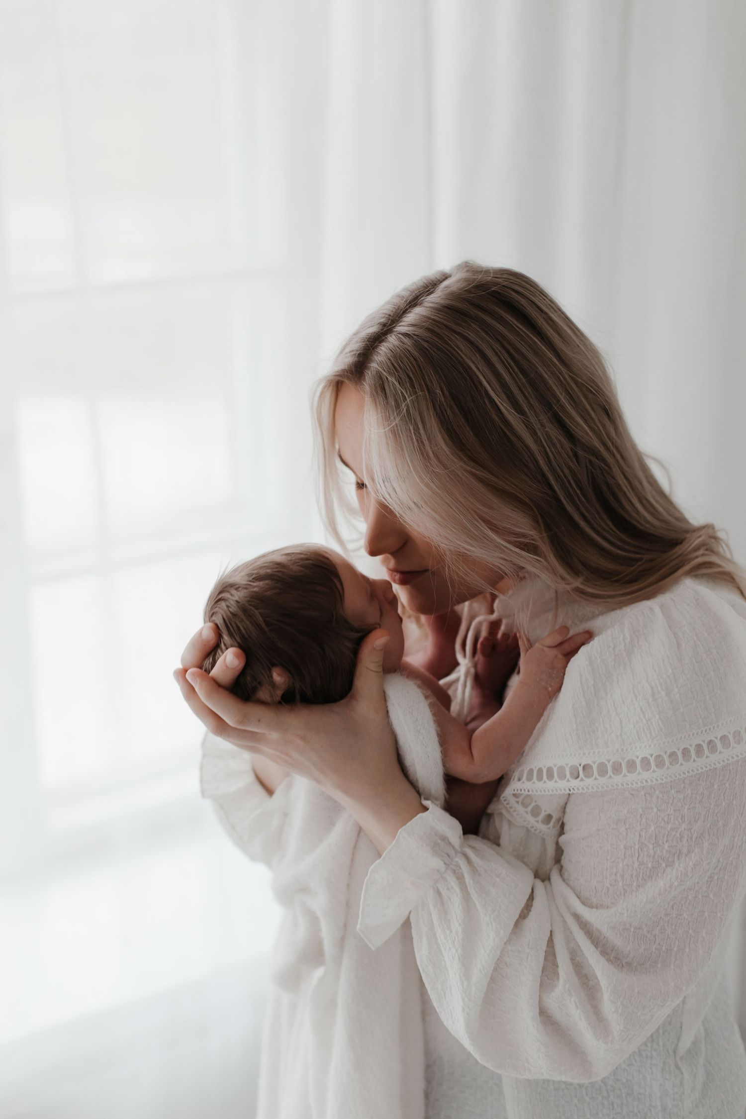 Intimate moment between mother and newborn baby in white room with natural window light.