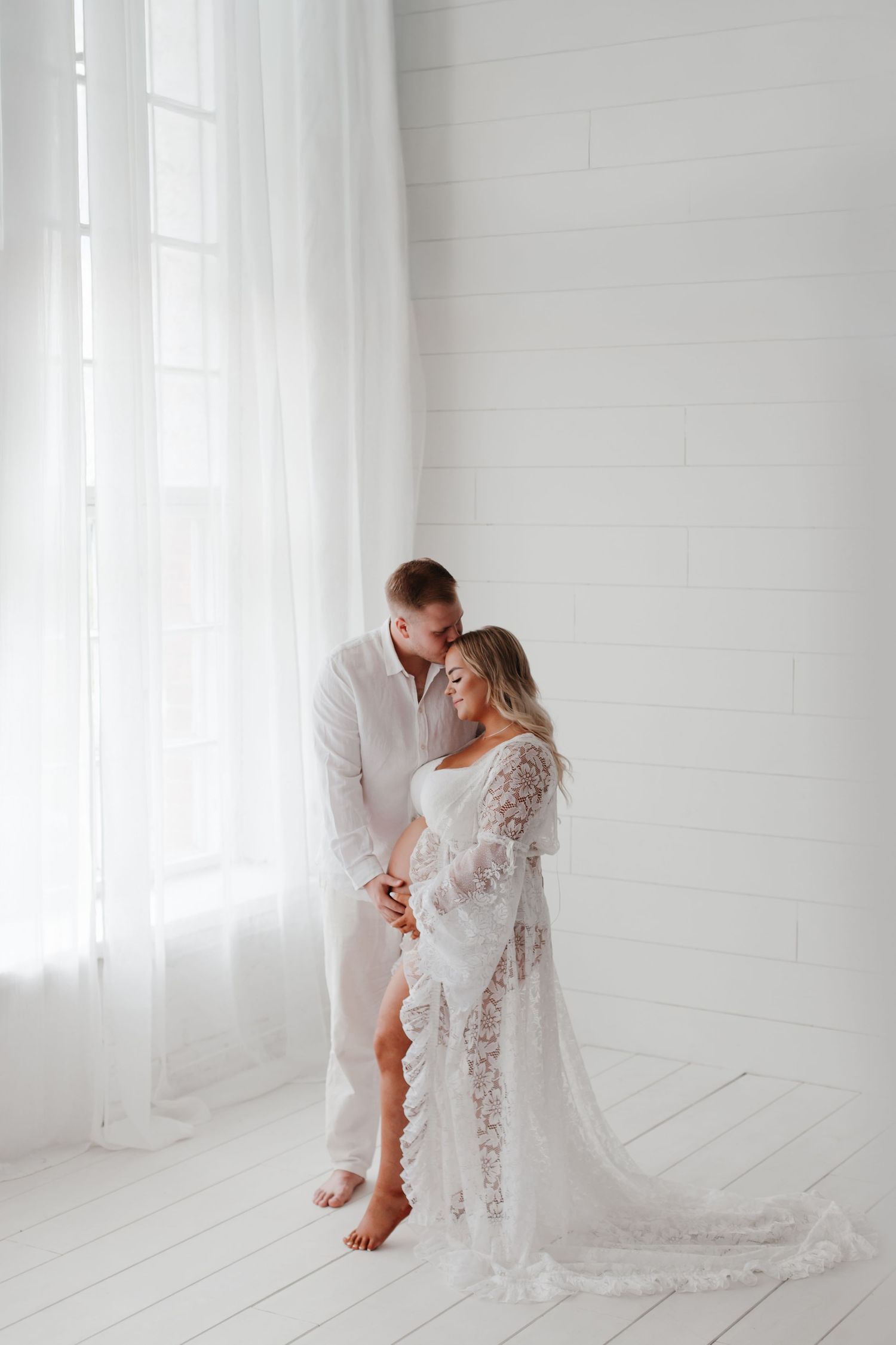 Ethereal maternity portraits in an all-white studio with delicate lace robe and romantic poses.