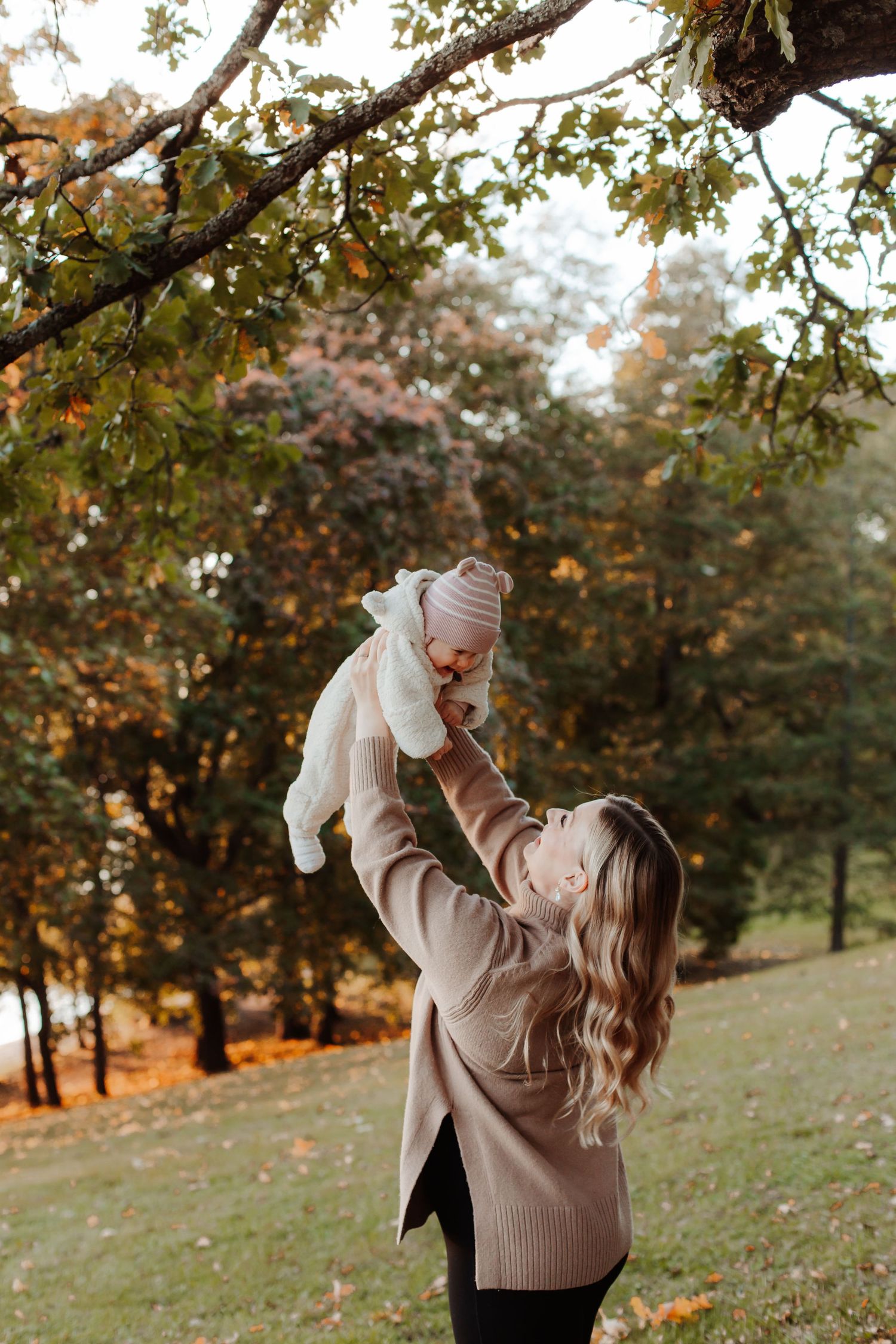 Person lifts baby playfully under autumn trees in park during golden hour.