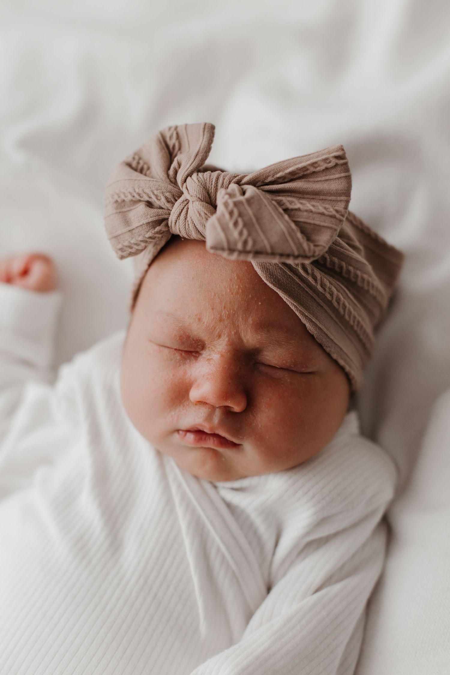 Newborn baby sleeping peacefully wearing white outfit and beige bow headband.
