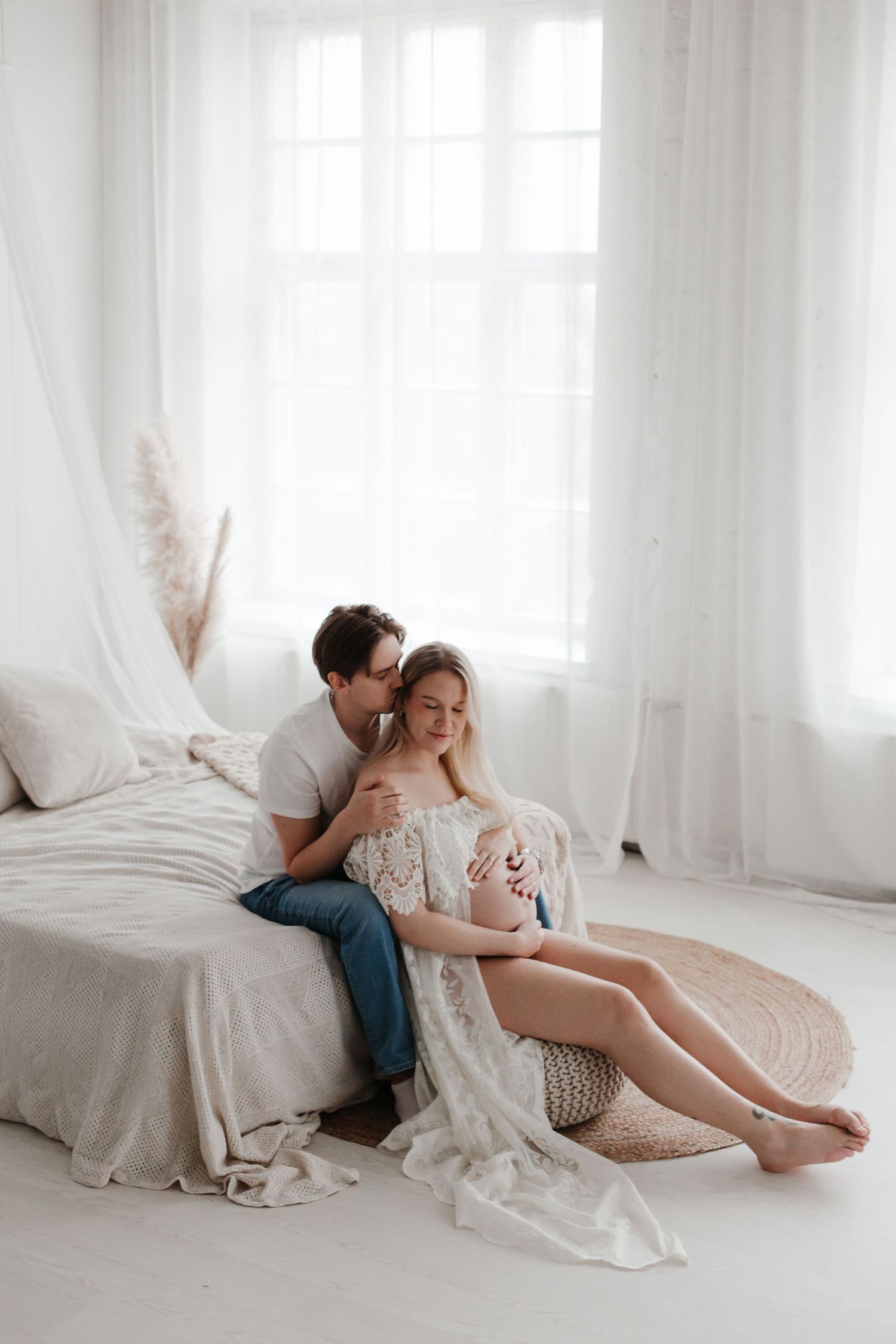 A romantic couple shares an intimate moment on white bedding in a bright room.