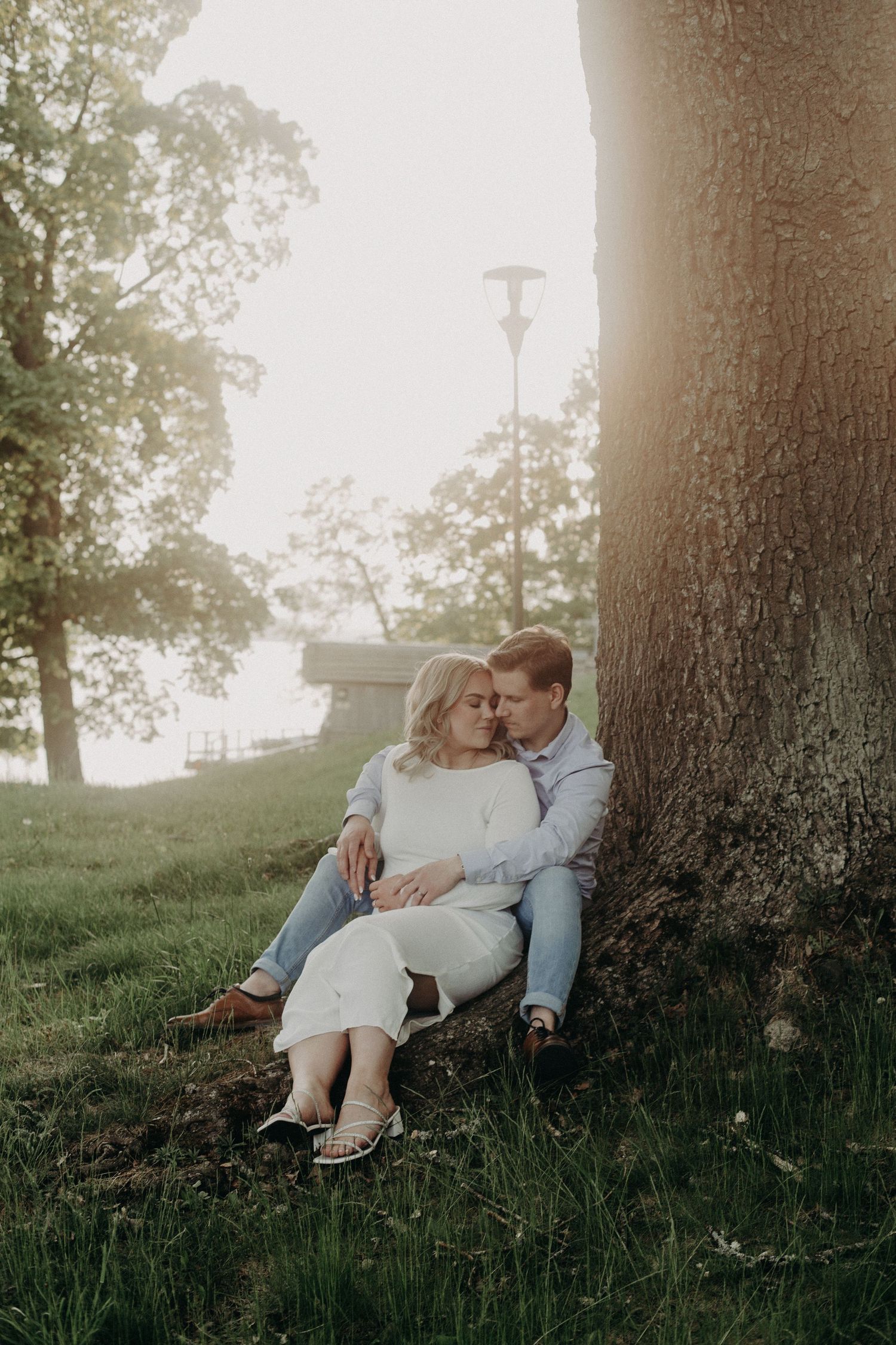 Couple sitting together against large tree trunk in park, sharing tender moment in warm sunset light.