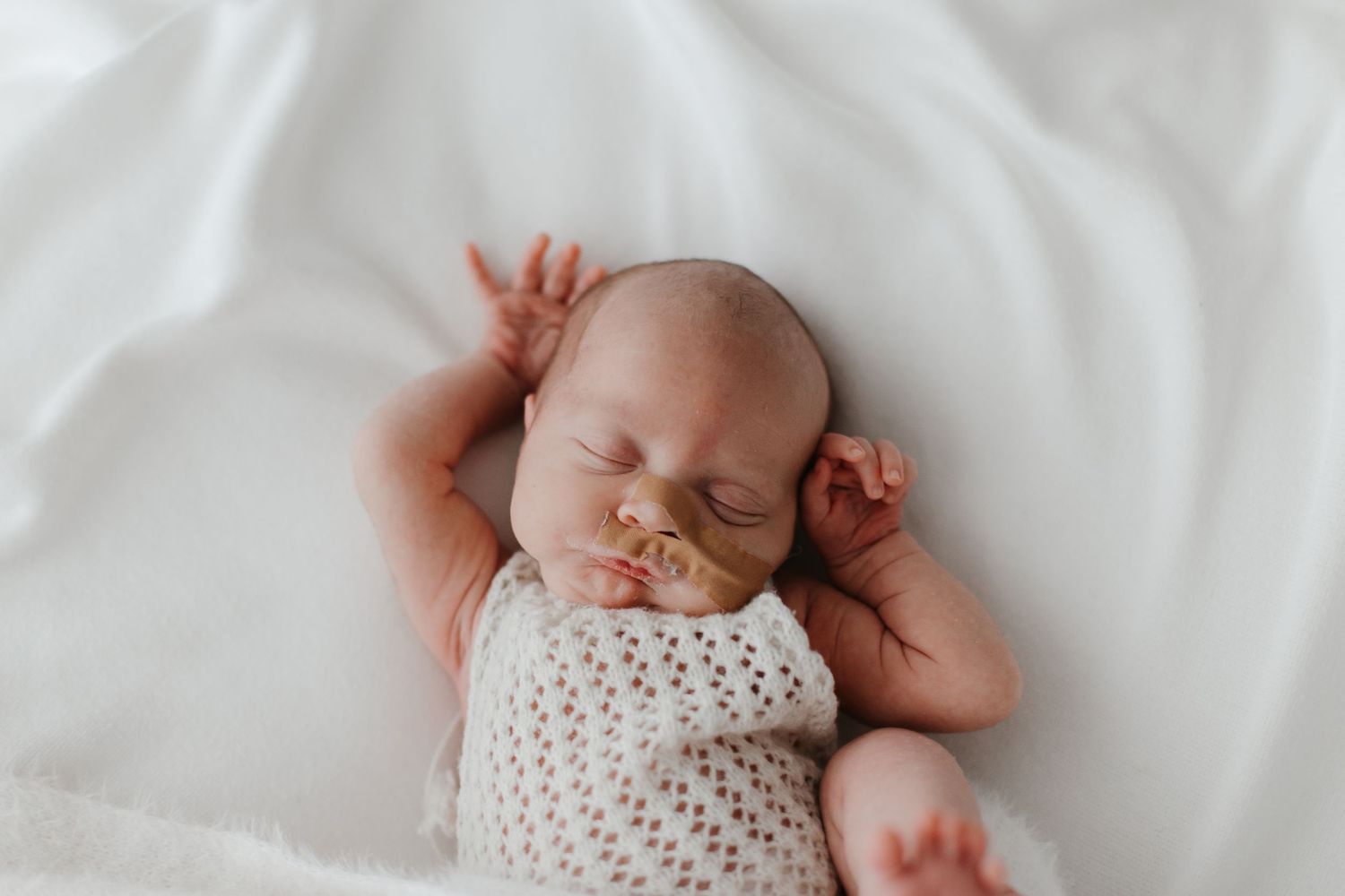 Sleeping newborn baby wearing white knit clothing against soft white bedding.