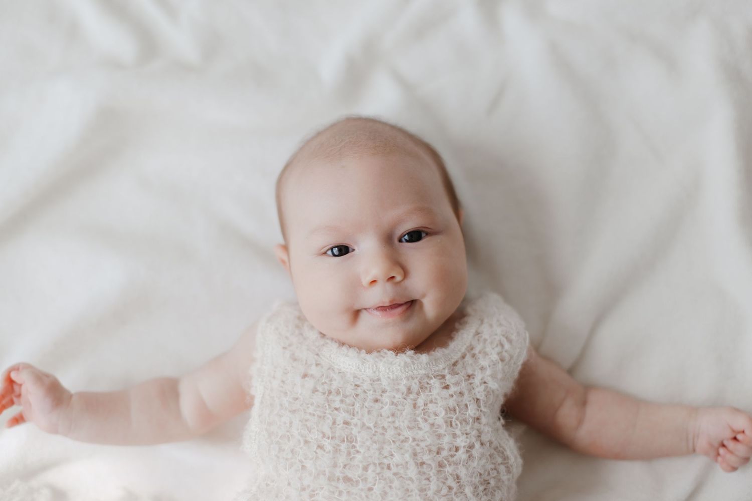 Smiling baby wearing cream knit romper lying on white bedding.