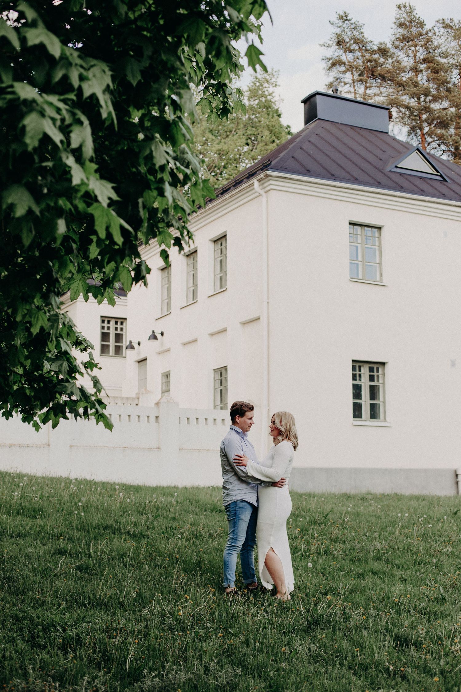 A couple embraces on green grass in front of a white manor house surrounded by lush trees.