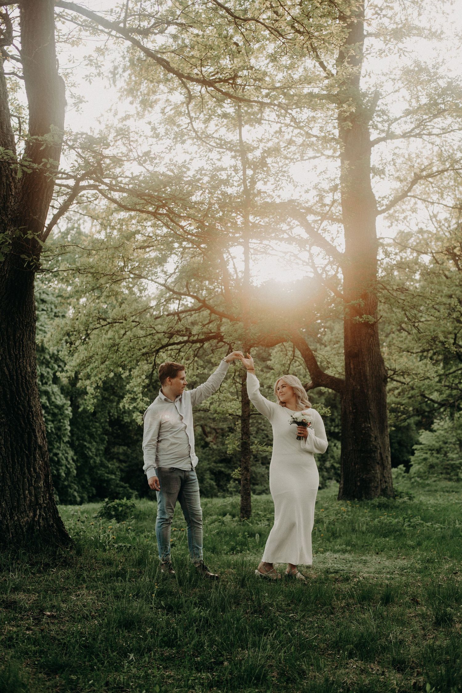 Couple dancing together in a sunlit forest clearing, bride in white dress holding bouquet, groom in casual attire.
