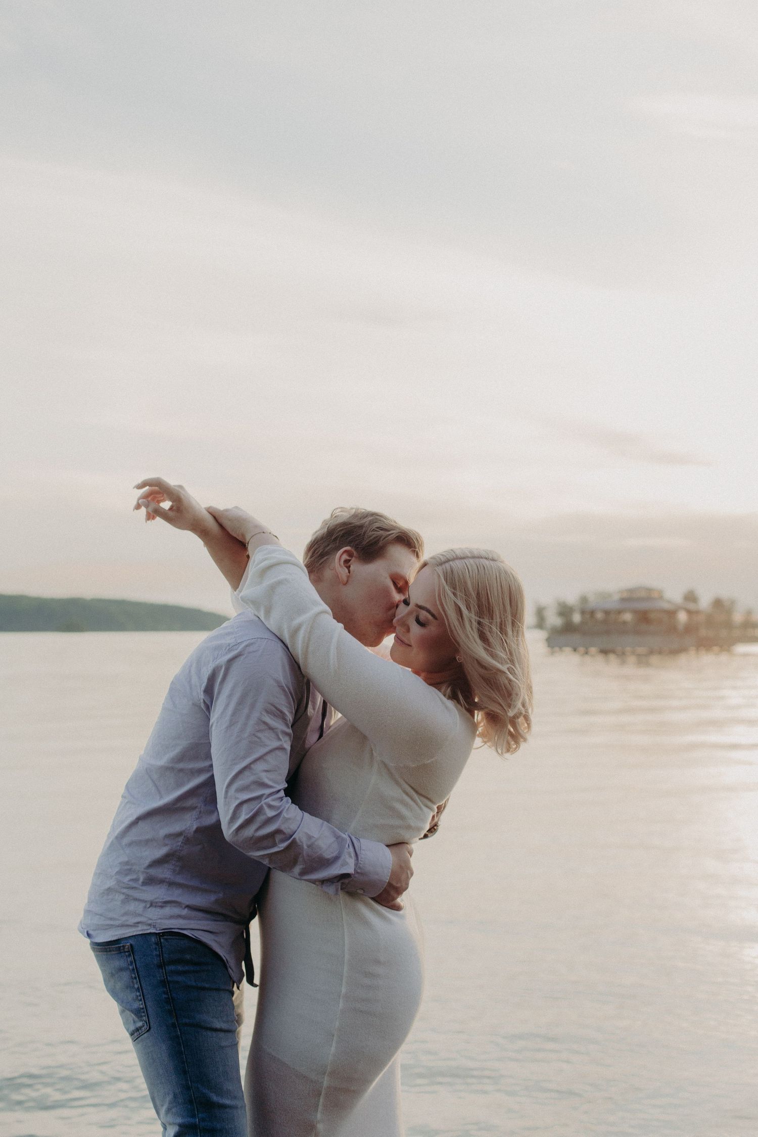 Romantic couple kissing and dancing by a serene lake at sunset, woman in elegant white fitted dress.
