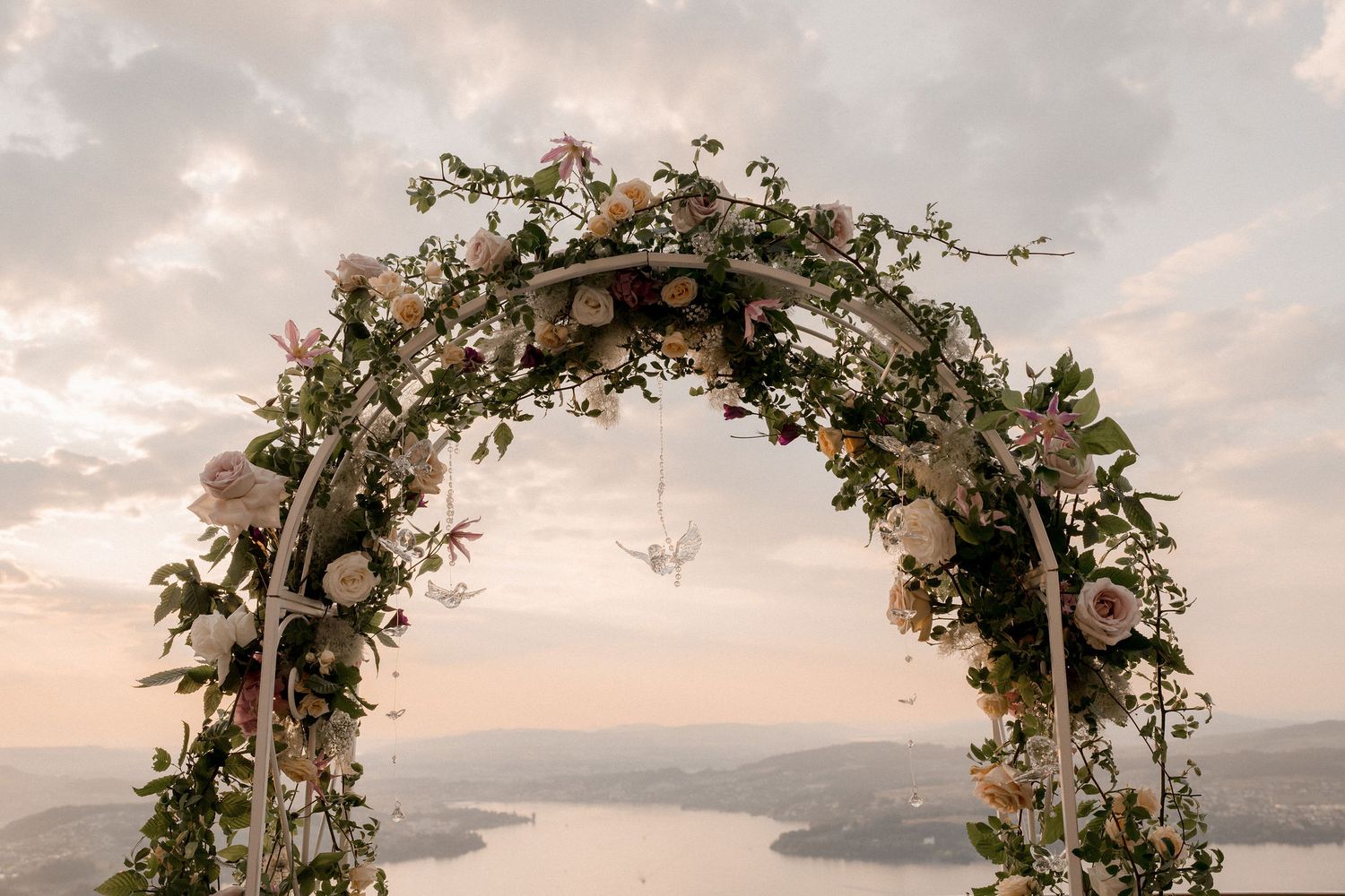 Floral wedding arch adorned with pink roses and greenery against a scenic lakeside sunset sky.