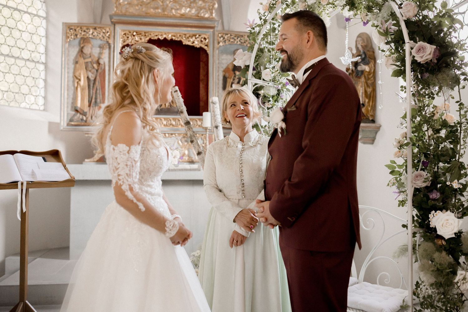 Bride and groom exchange vows at a floral church altar with an officiant in a white dress.