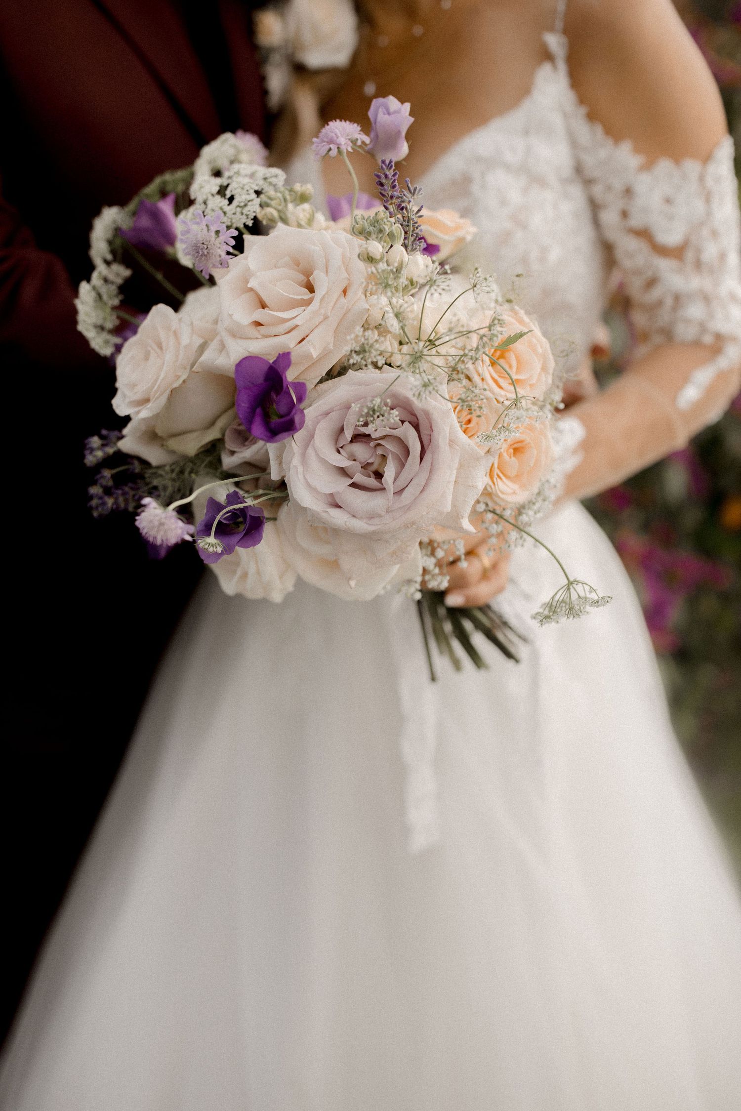 Bride holding a romantic bouquet of blush, lavender roses and purple flowers in a white lace wedding gown.