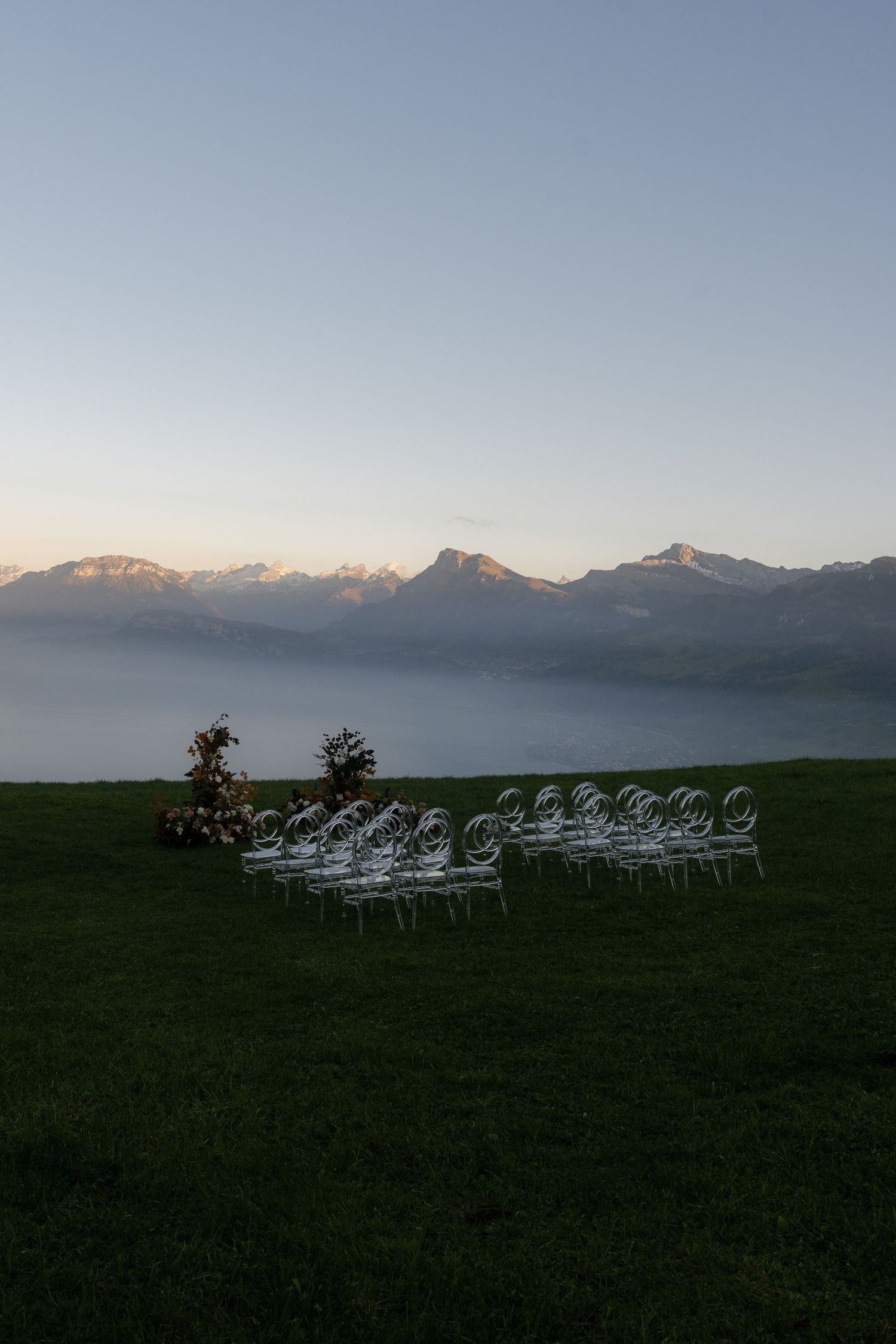 Outdoor wedding ceremony setup with white chairs on green grass, misty mountain backdrop at dusk.
