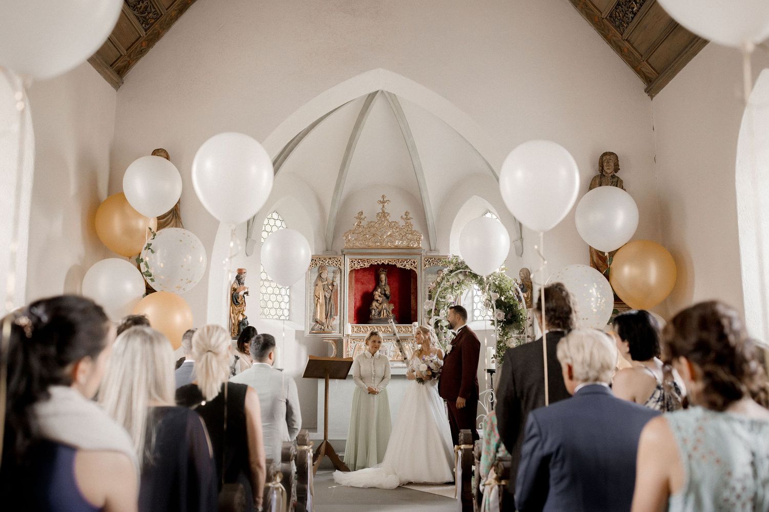 Elegant church wedding ceremony with large white and gold balloons, guests seated in pews, bride and groom at the altar.