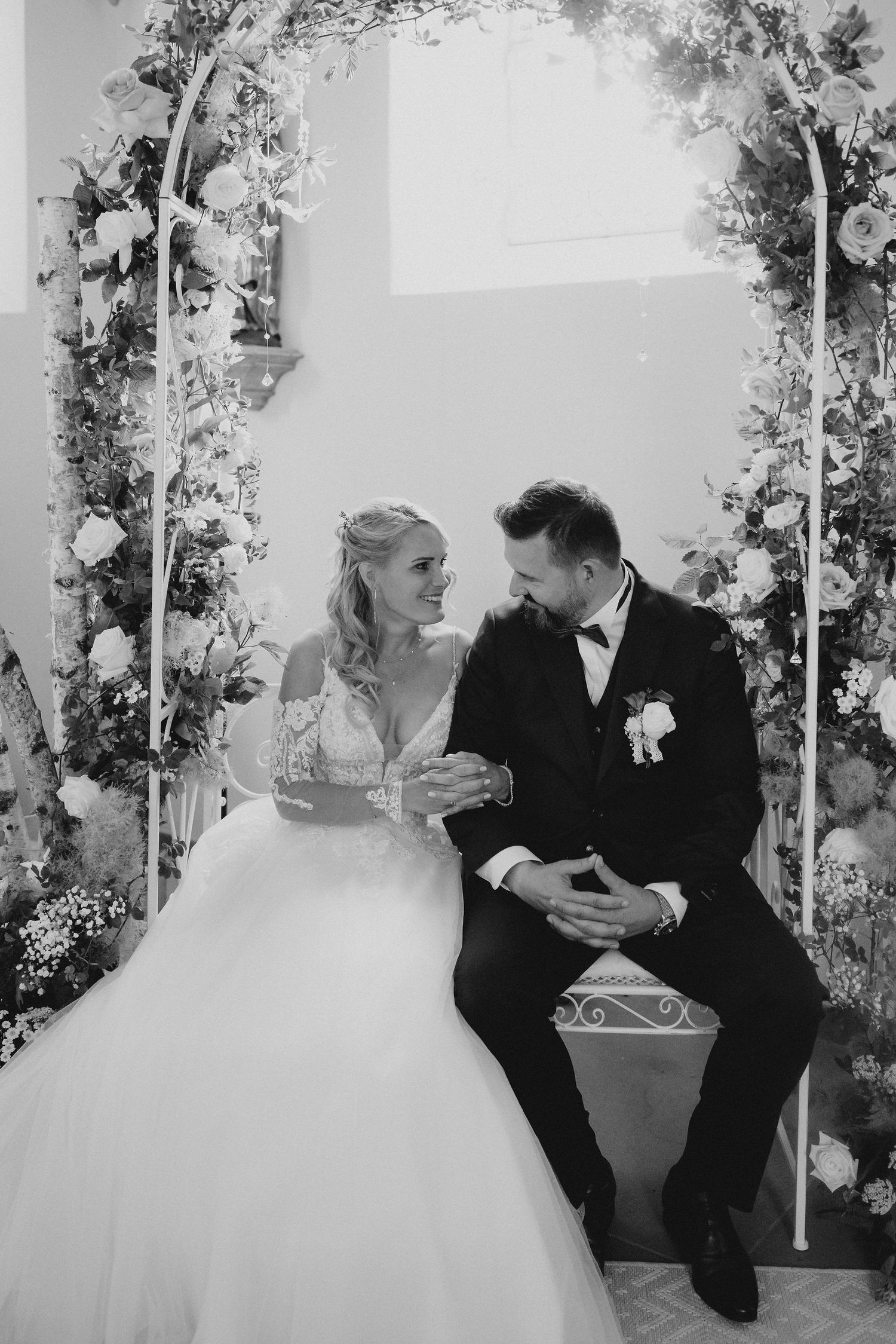Black and white wedding photo of bride and groom sitting under floral arch, gazing lovingly at each other.