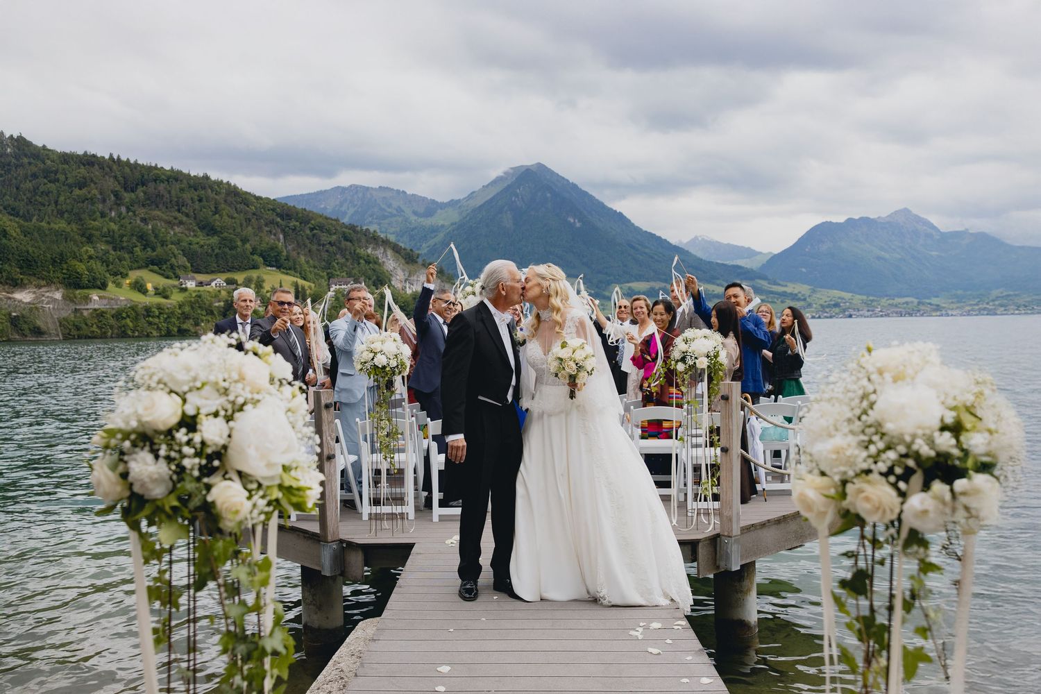 Romantic outdoor wedding ceremony on a lakeside dock with mountain views, white floral arrangements, and guests cheering the newlyweds.