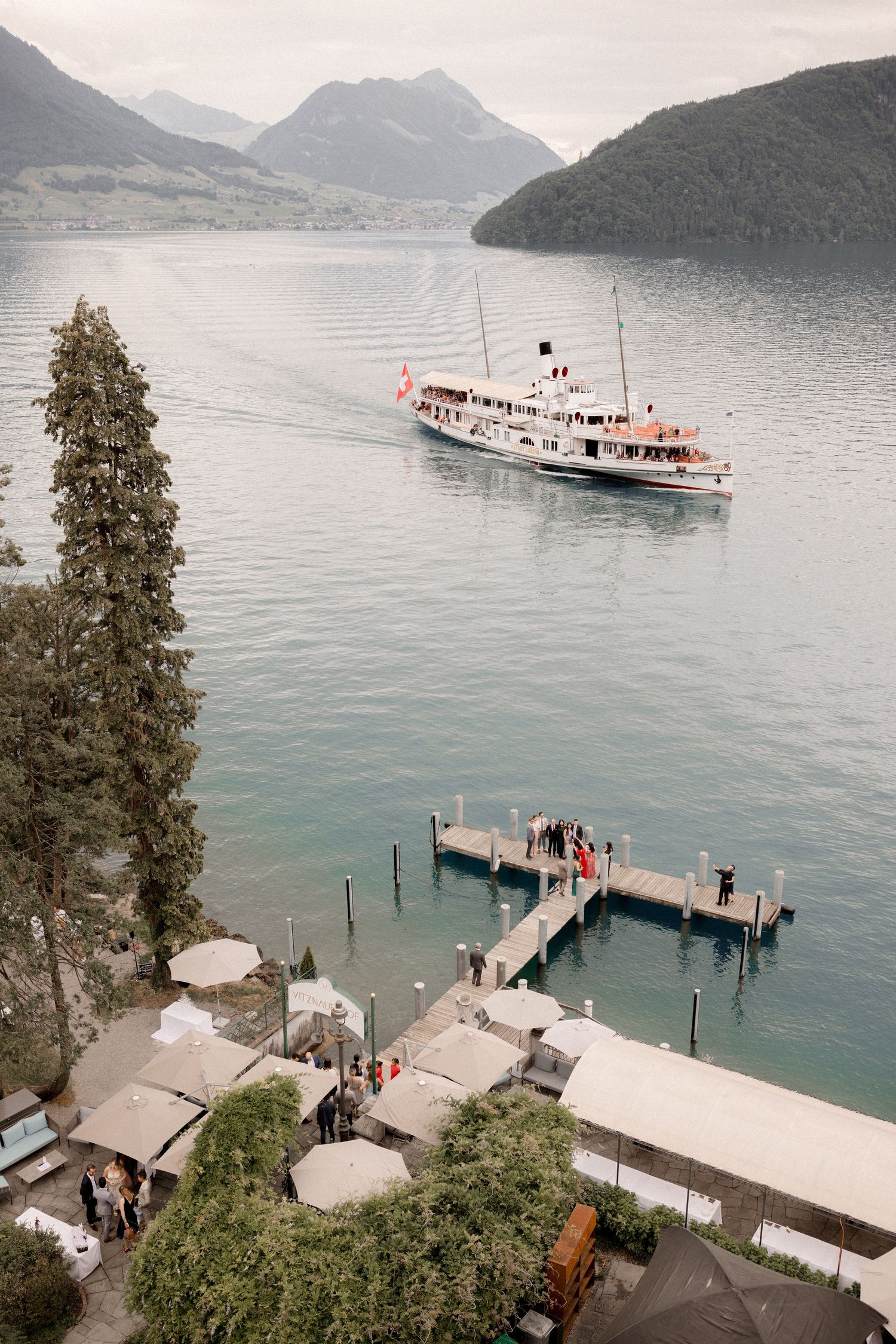 A vintage passenger ship cruises across a mountain lake with a wooden dock in the foreground.